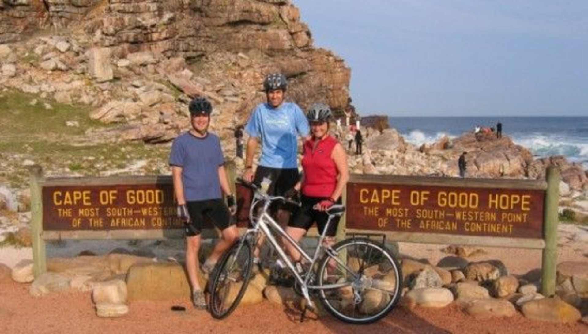The image shows three people standing next to a bicycle in front of a sign indicating the Cape of Good Hope, with a rocky coastline and ocean visible in the background.