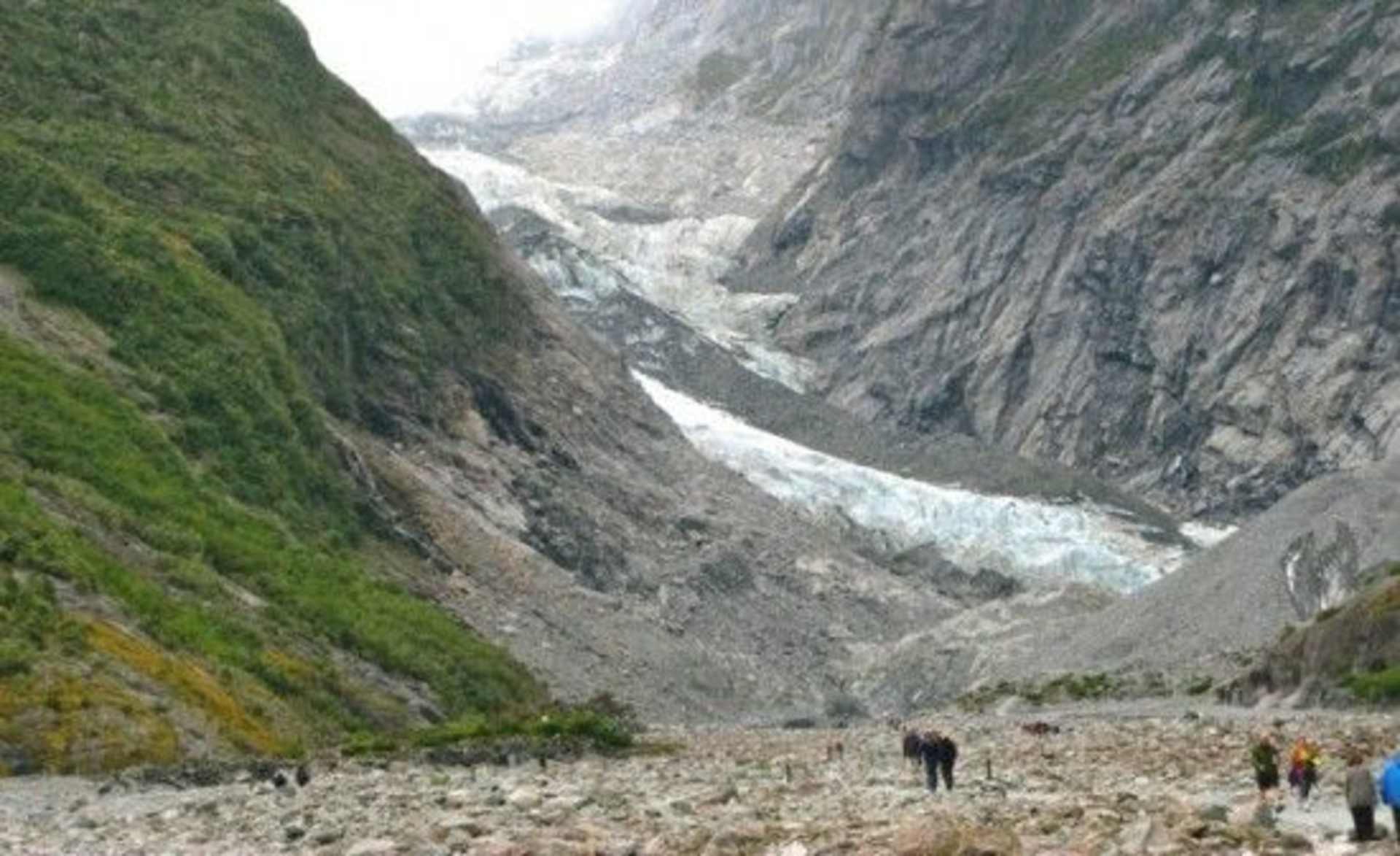A rugged, mountainous landscape with a glacier visible in the distance, and several people walking along a path in the foreground.
