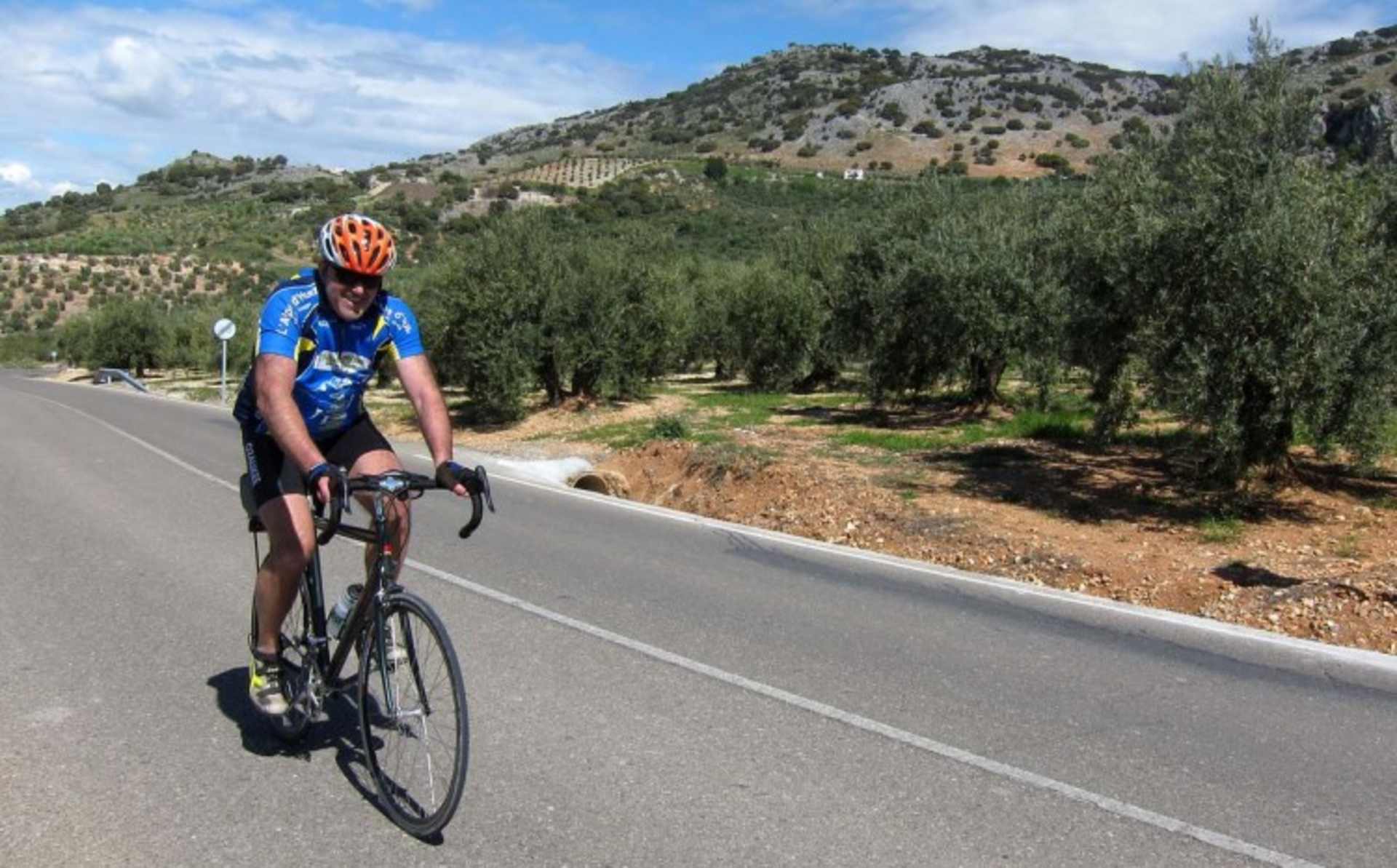 A cyclist in a blue jersey and helmet rides along a paved road surrounded by rolling hills, olive groves, and a clear blue sky.