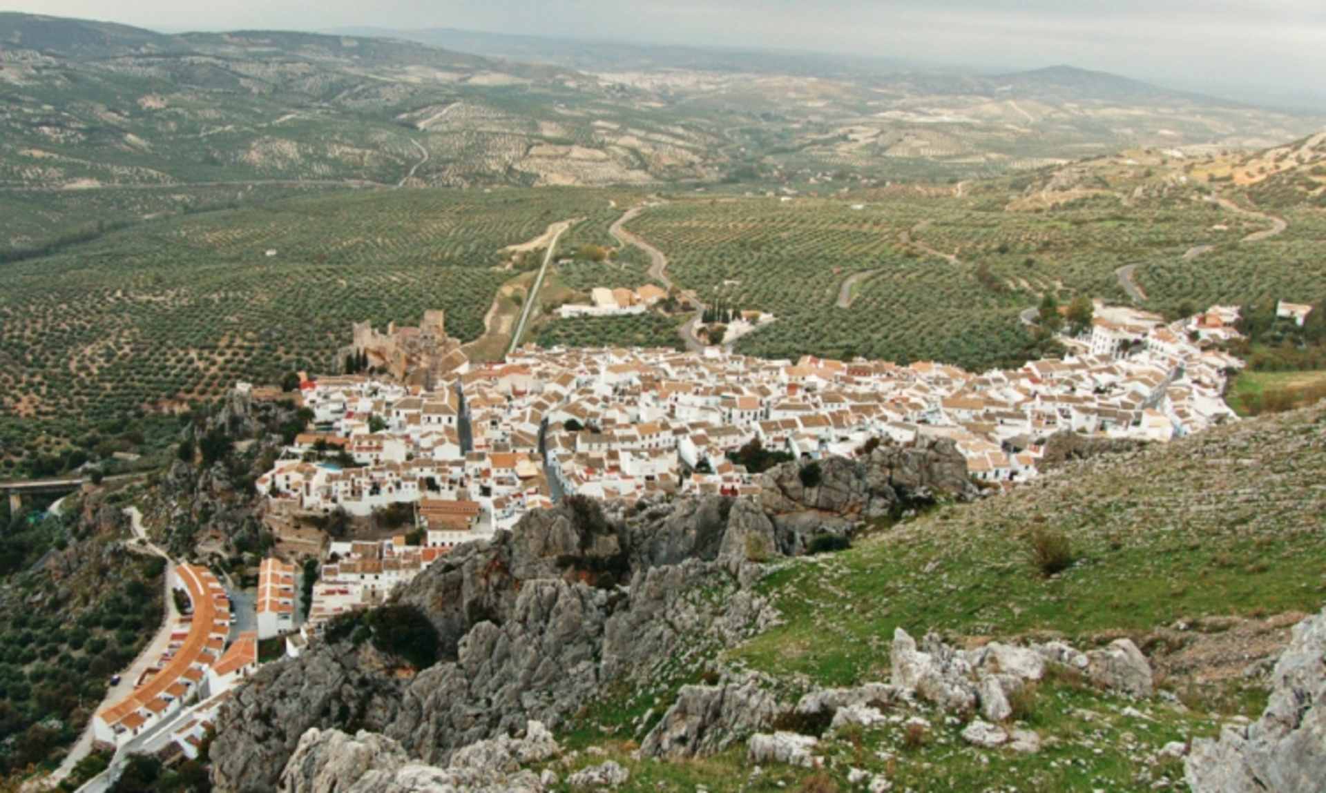 A picturesque town nestled among rugged mountains, with winding roads and buildings cascading down the steep slopes, surrounded by lush greenery and a vast, hilly landscape.