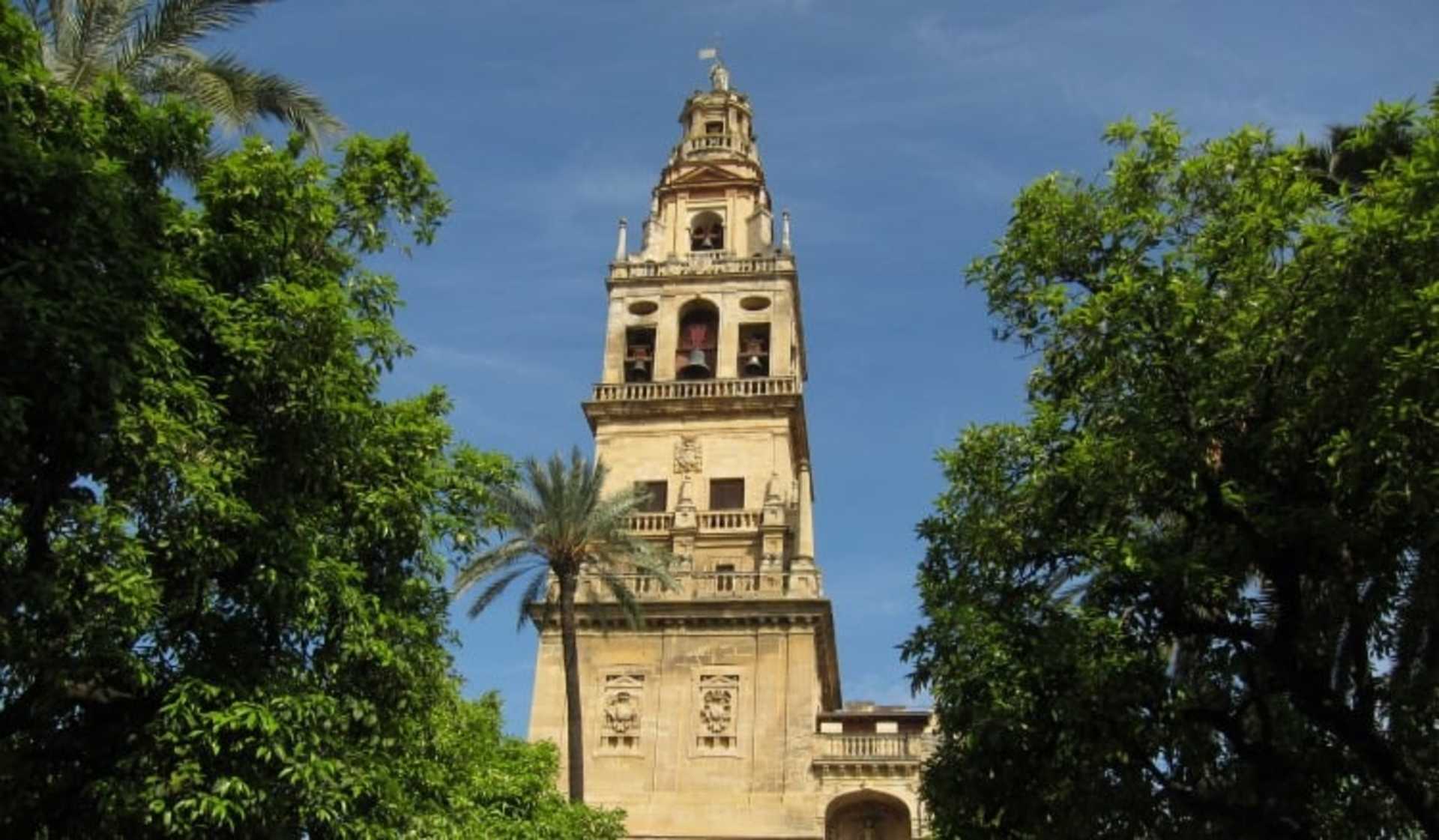 A tall, ornate bell tower rises above the lush, green foliage in the foreground, set against a clear blue sky.