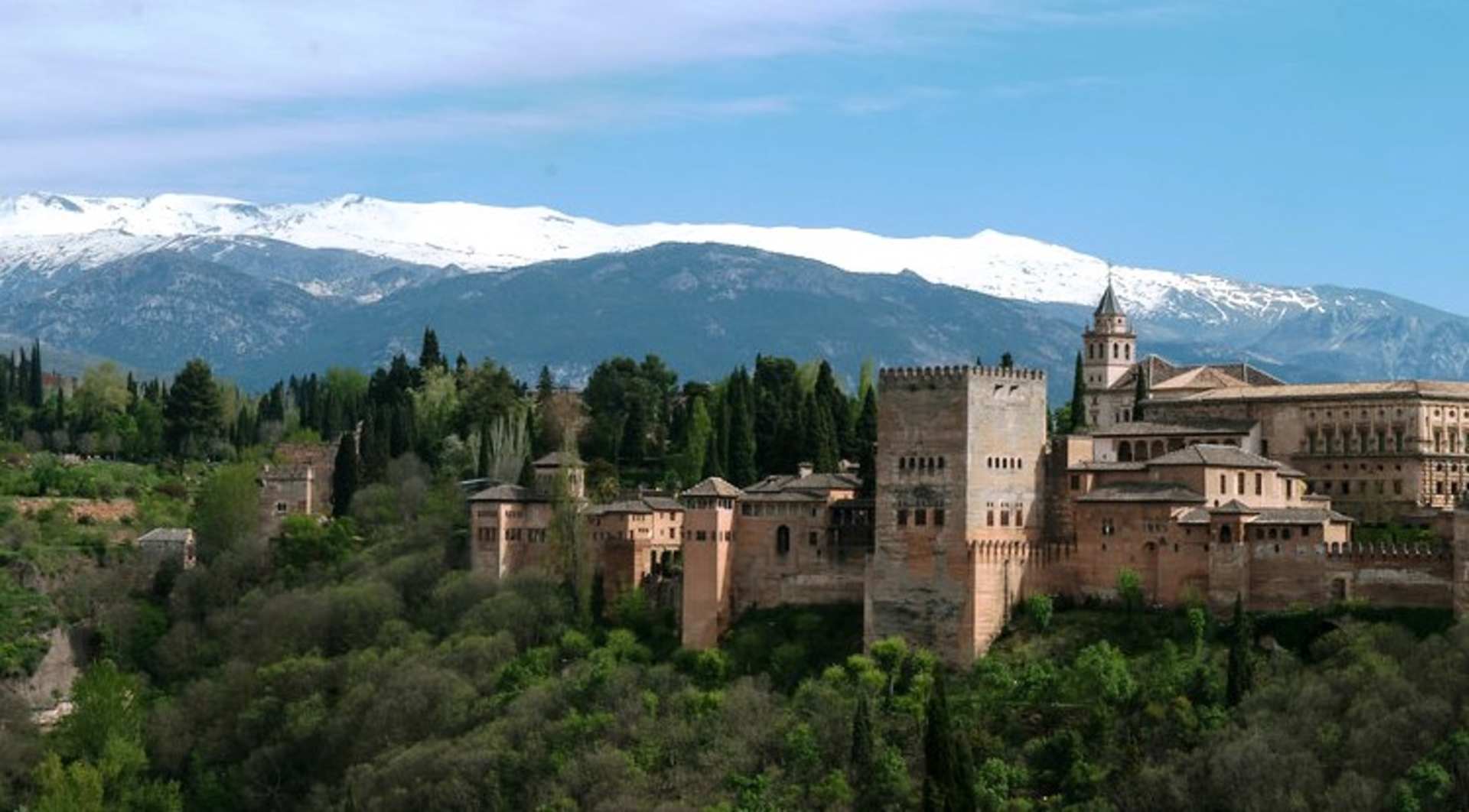 A picturesque medieval castle nestled among lush greenery, with snow-capped mountains towering in the background under a clear blue sky.