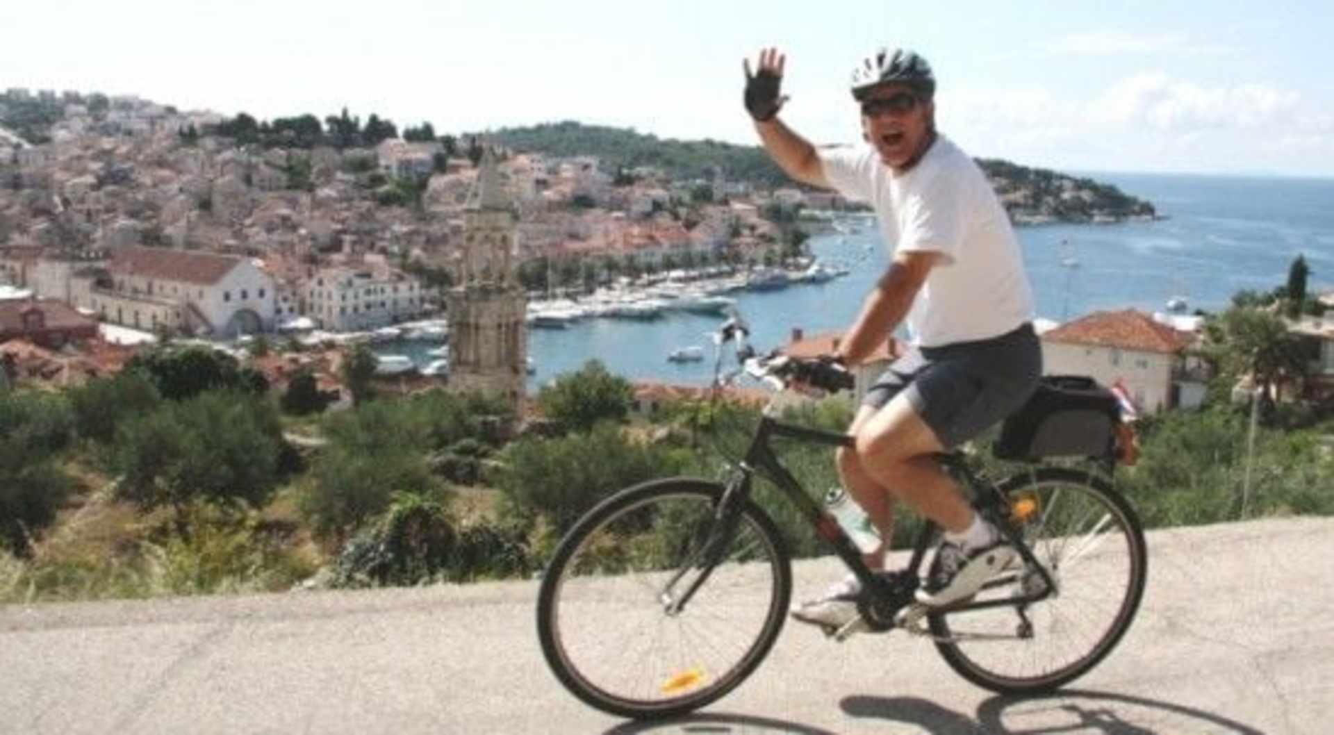 A person on a bicycle is riding on a road overlooking a picturesque coastal town with colorful buildings and a harbor in the background.