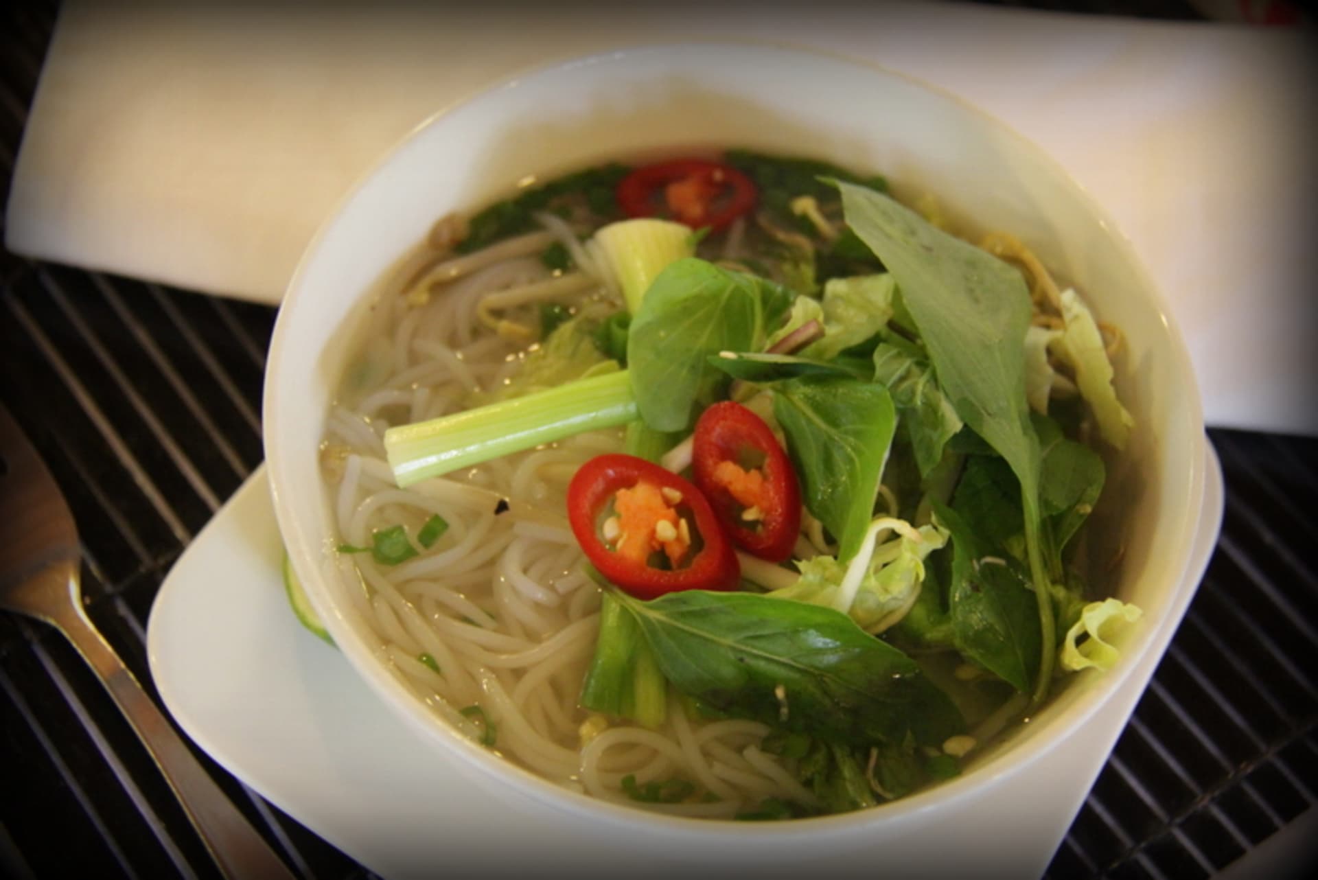 A white bowl filled with noodles, vegetables, and a spicy broth, set against a dark background with a pair of chopsticks.