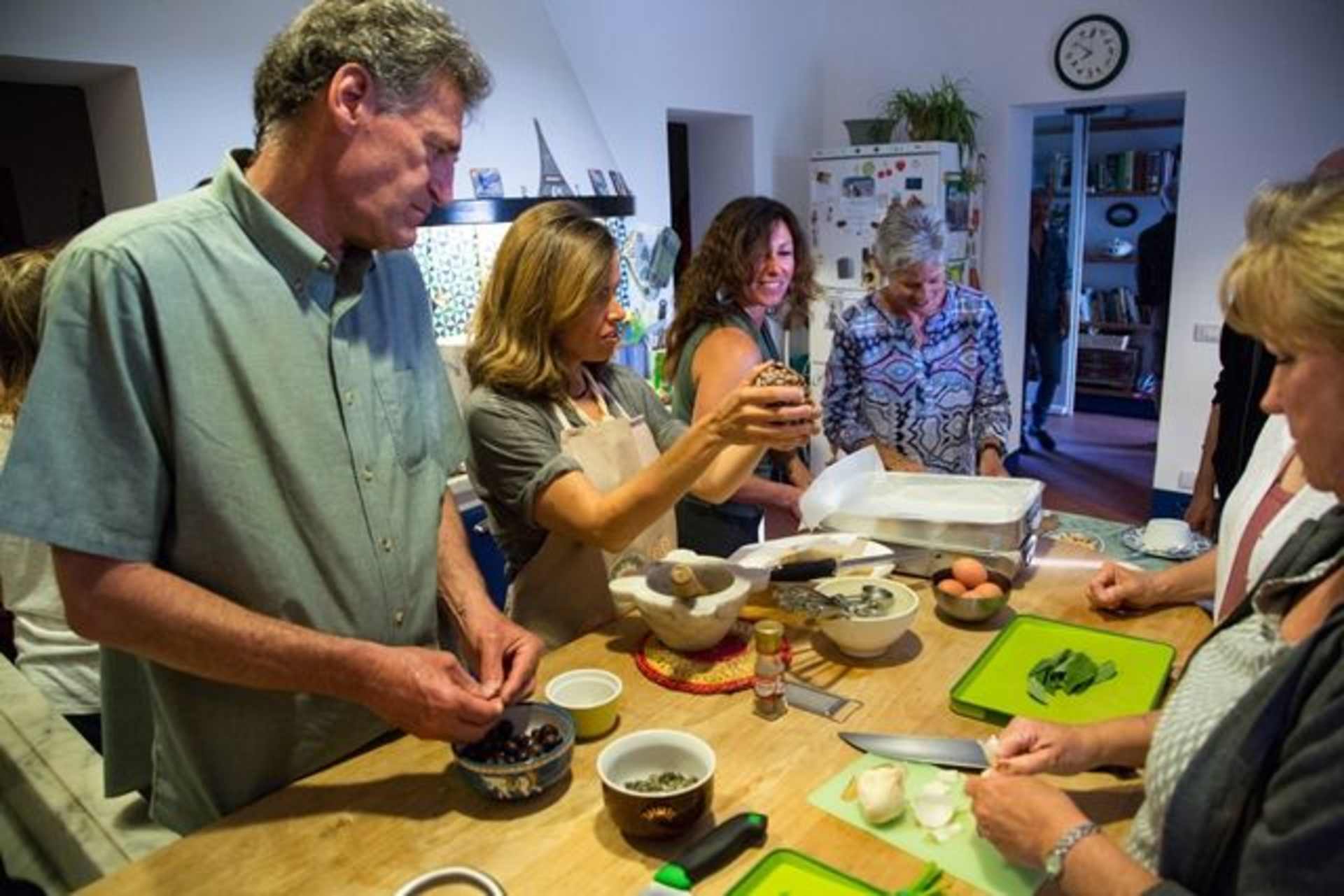 A group of people gathered in a kitchen, engaged in what appears to be a cooking or food preparation activity, with various food items and utensils visible on the table.