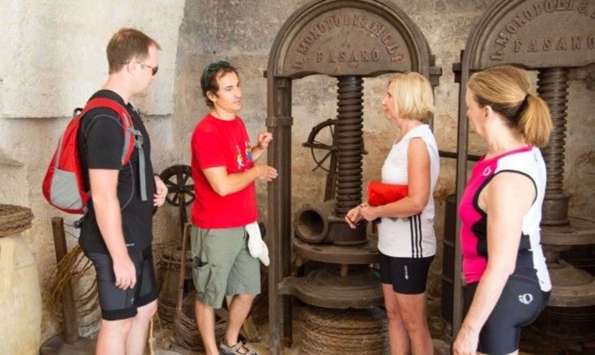 Four people, two men and two women, are standing in what appears to be an old industrial or workshop setting, with large machinery and equipment visible in the background.