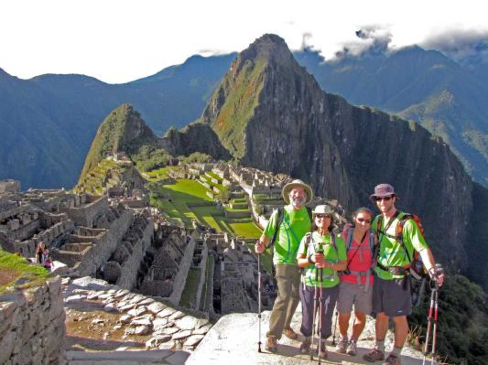 A group of hikers stands on a stone pathway overlooking the ancient Inca ruins of Machu Picchu, with the majestic Andes mountains rising in the background.