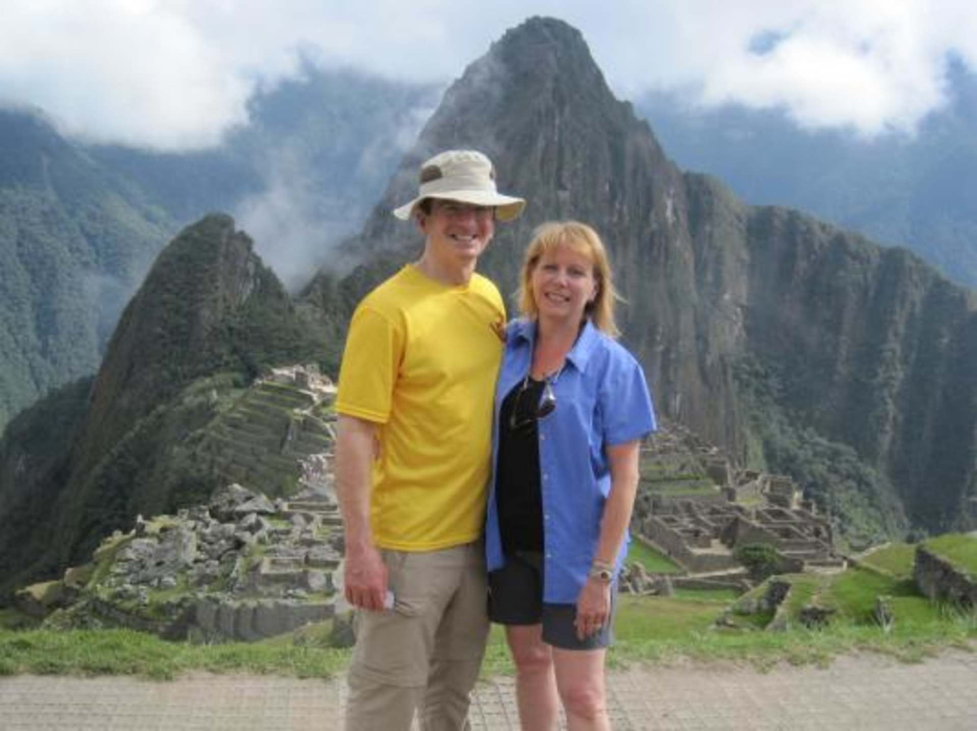 The image shows two people, a man and a woman, standing in front of the ancient Inca ruins of Machu Picchu, with the iconic mountain peaks of the Andes in the background.