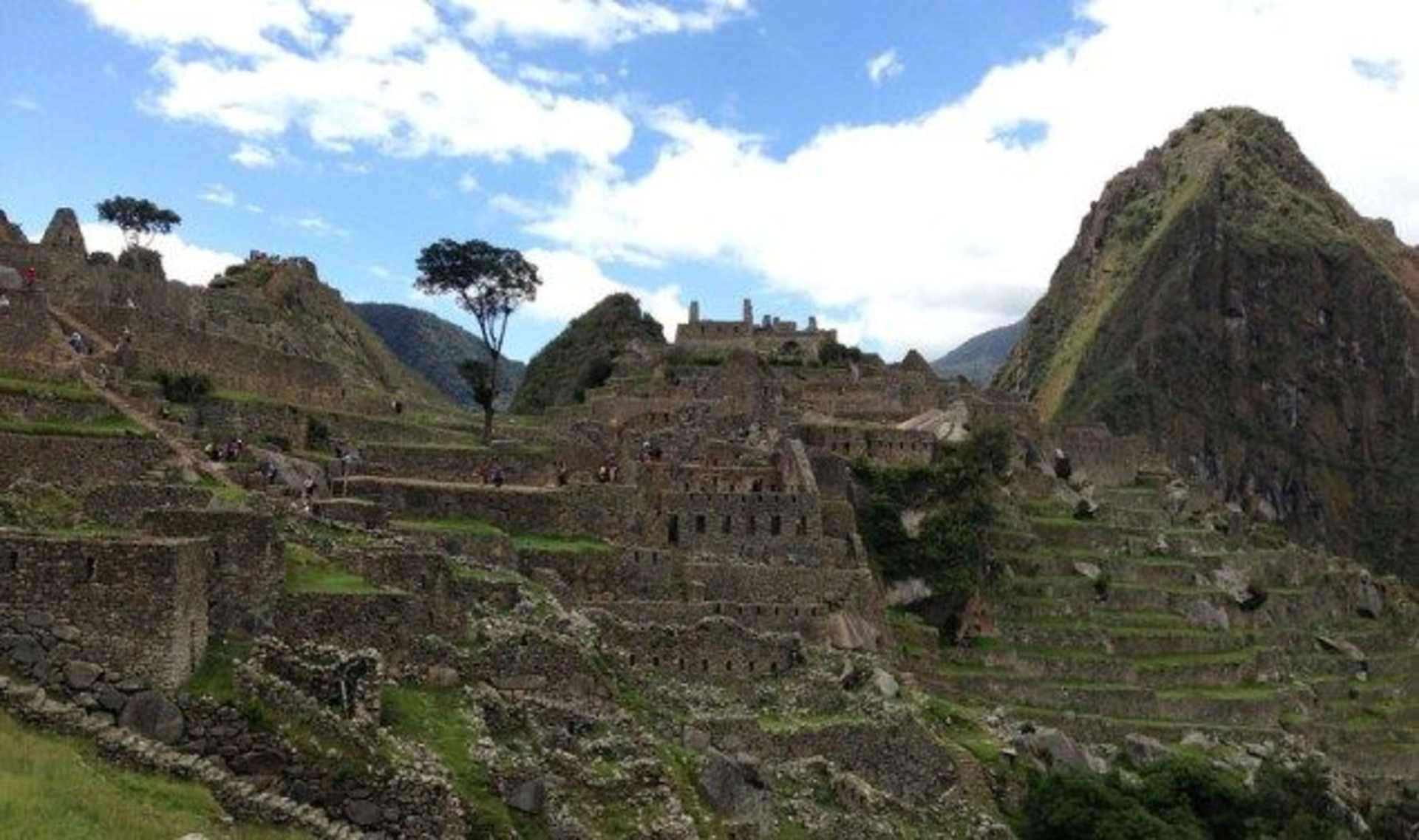 Terraced stone structures nestled among towering mountains, with a lush, verdant landscape and a bright, cloudy sky in the background.