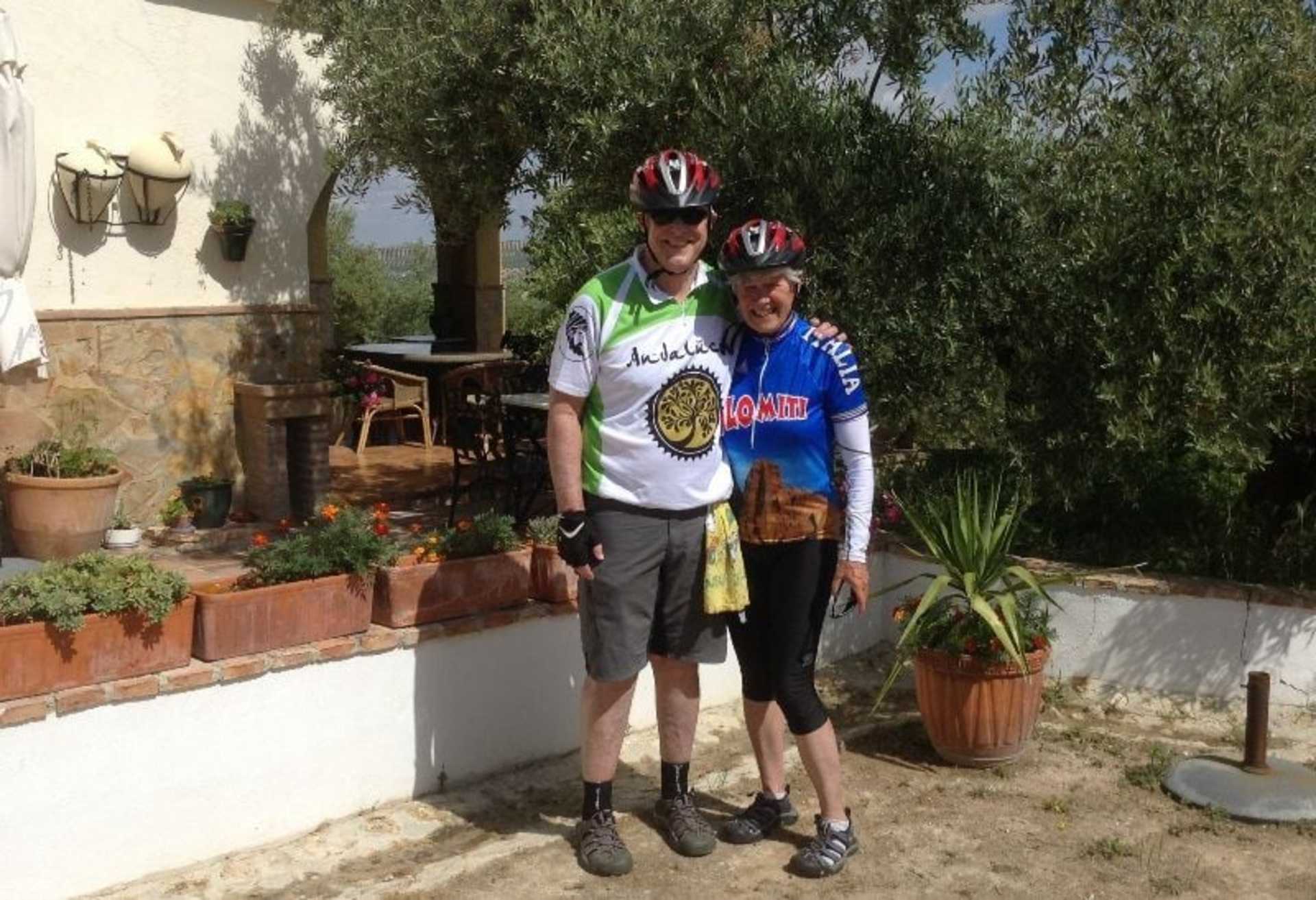 Two people, a man and a woman, are standing in a garden-like setting with potted plants and a wall in the background. They are wearing cycling gear, including helmets, and appear to be taking a break from a bike ride.