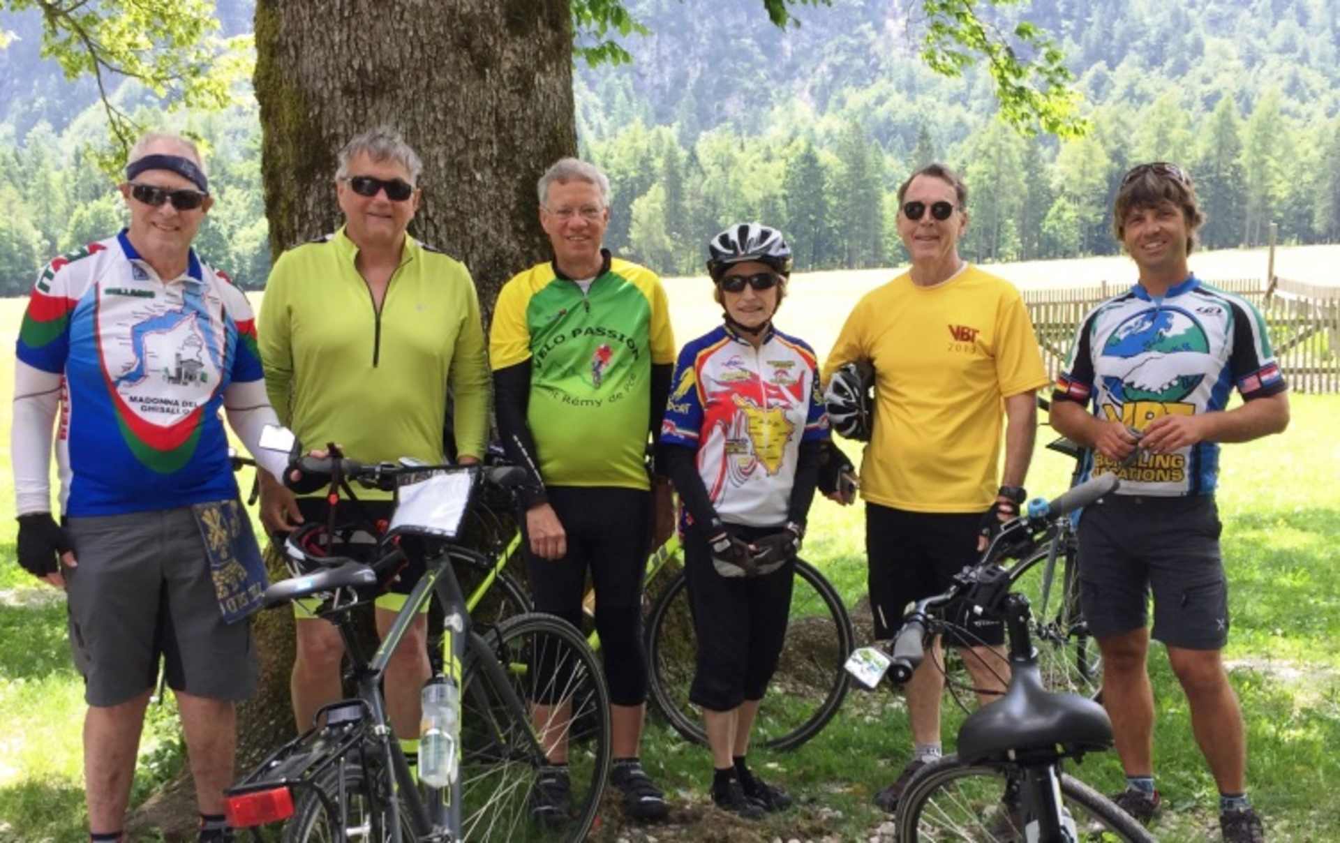 A group of cyclists, dressed in colorful cycling gear, stand together in a scenic outdoor setting surrounded by lush greenery and trees.