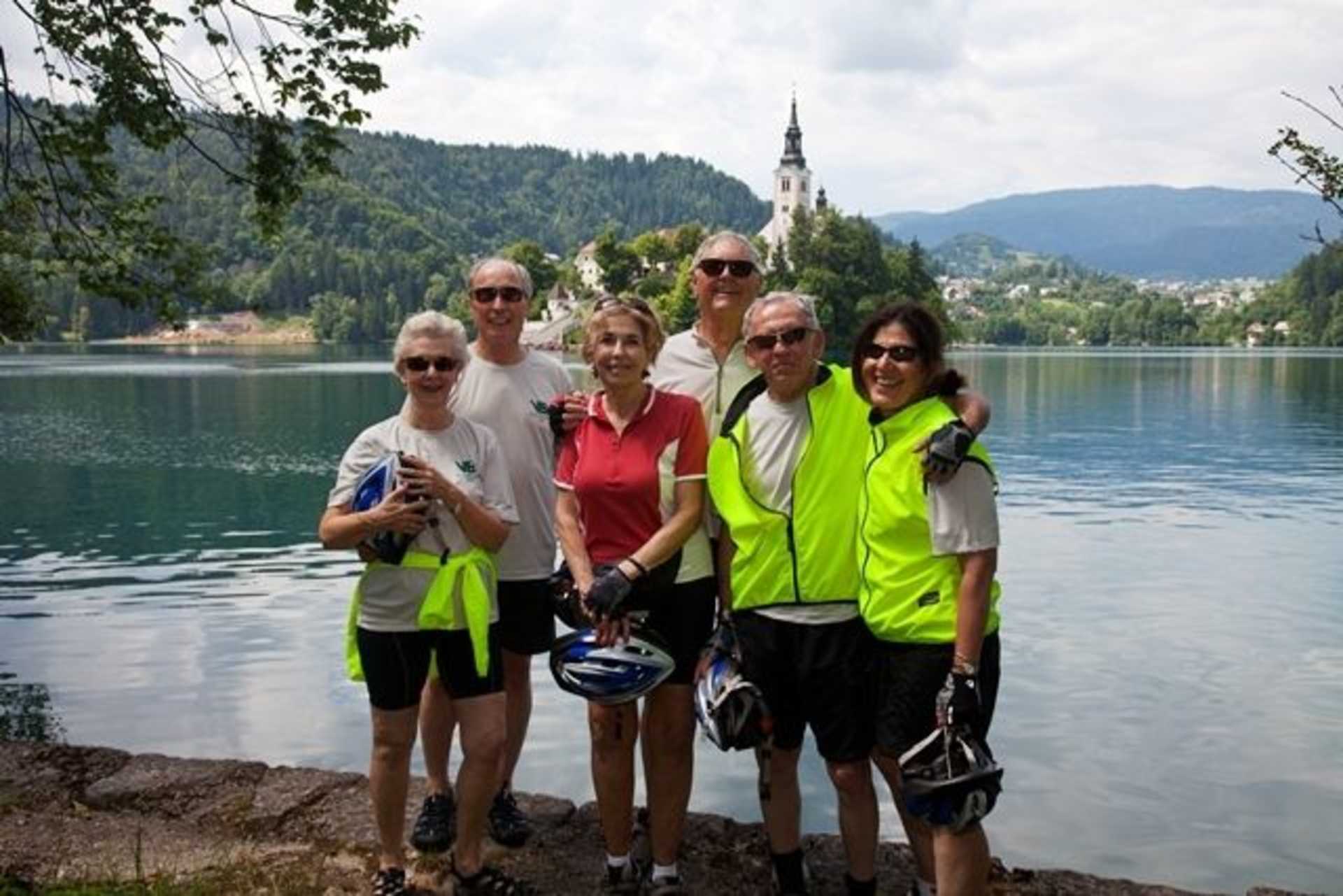 A group of people posing for a photo in front of a scenic lake with a church in the background, surrounded by lush greenery and mountains.