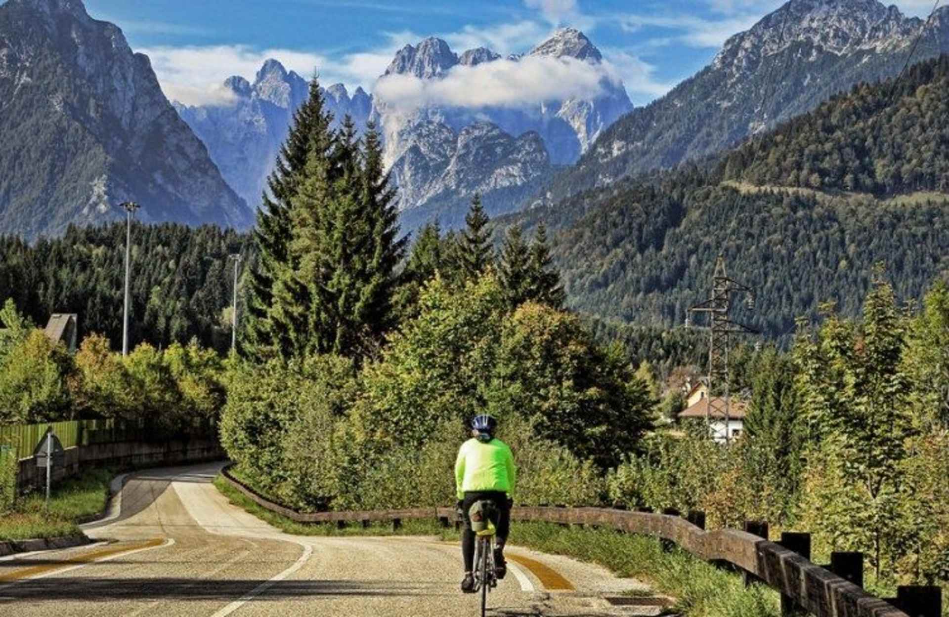 A scenic mountain landscape with a cyclist riding on a paved path surrounded by lush green forests and towering peaks in the background.
