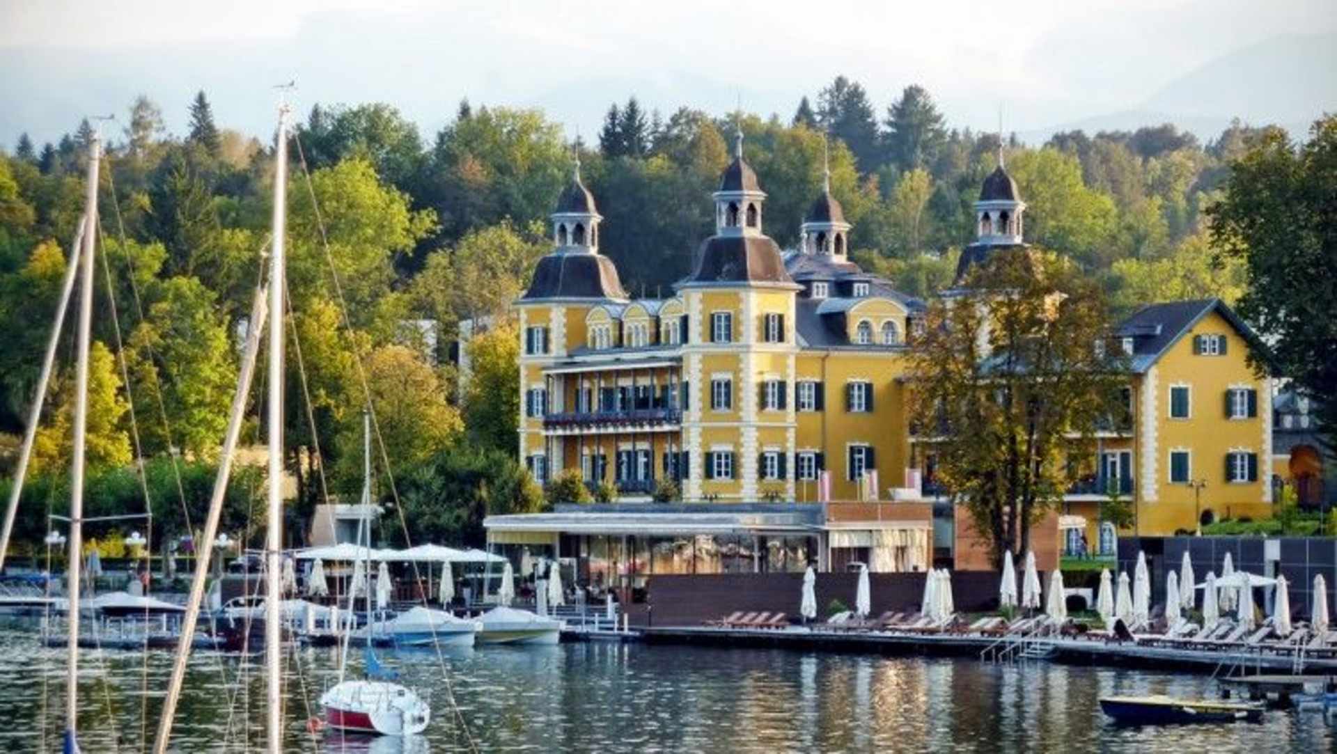 A picturesque harbor with sailboats and yachts moored in the foreground, surrounded by a charming, ornate yellow building with towers and a lush, forested backdrop.