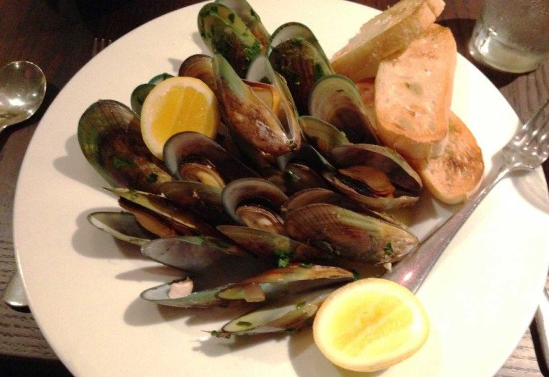 A plate filled with steamed mussels, accompanied by slices of lemon and a piece of bread, set against a dark background.