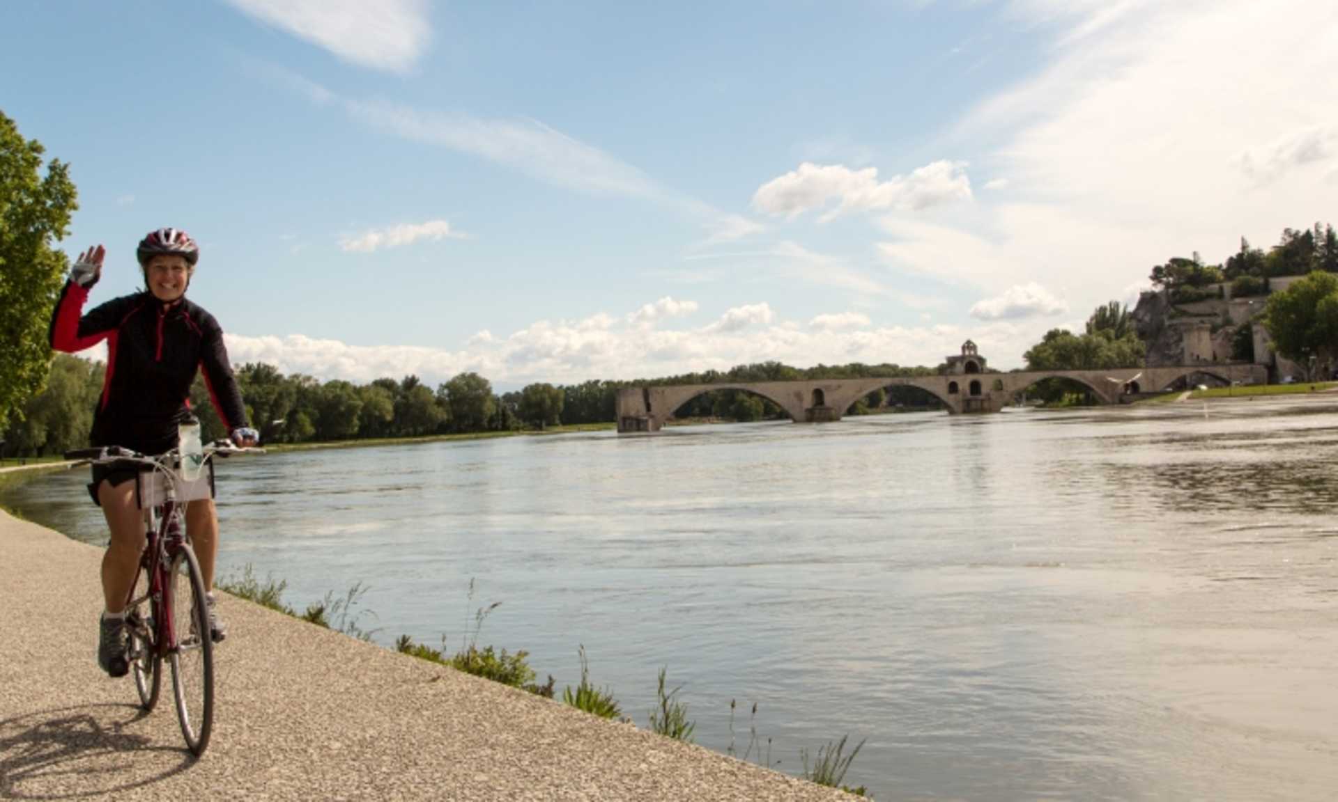 A cyclist rides along the riverbank, with a historic stone bridge and scenic landscape visible in the background.