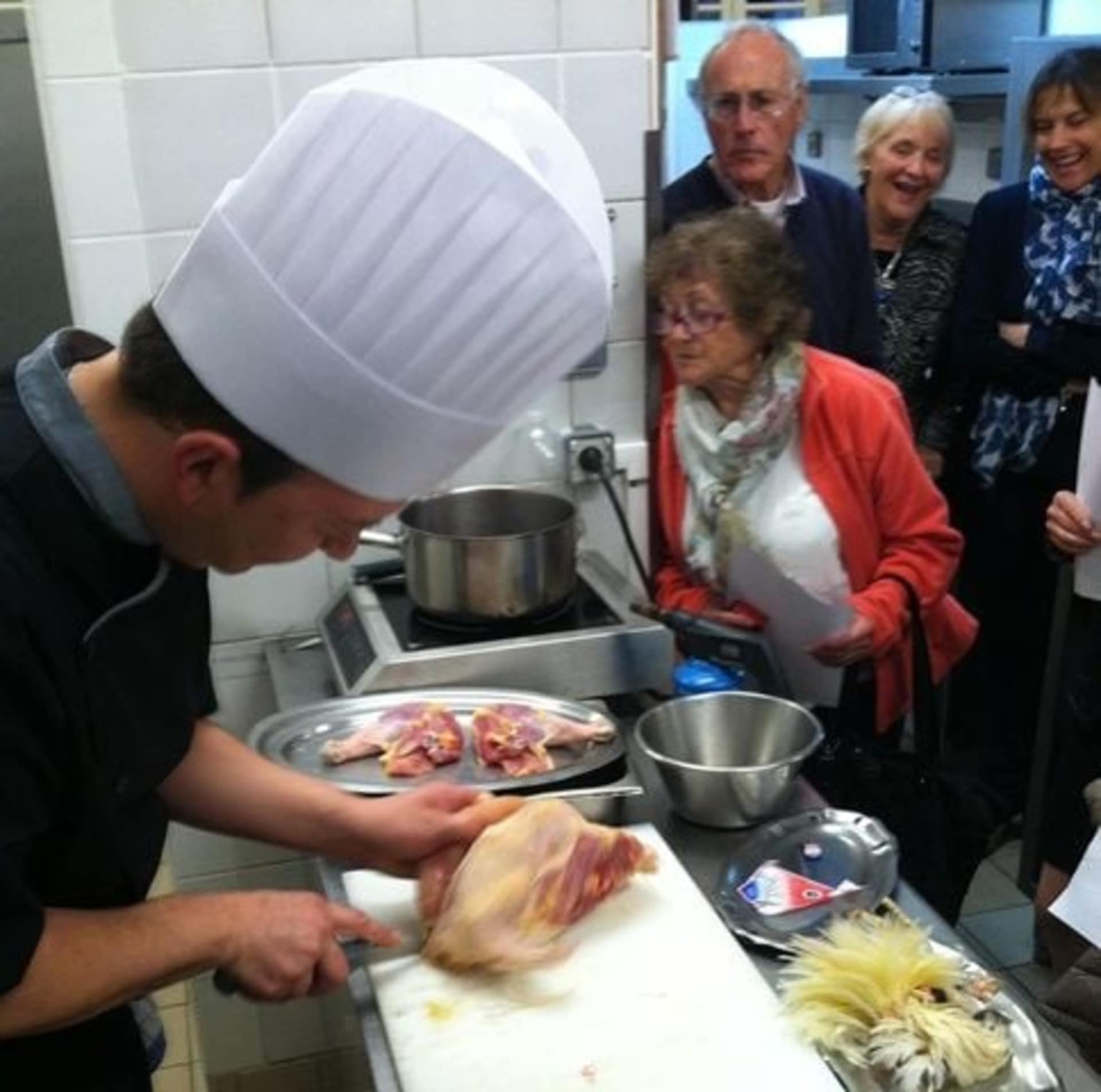 A chef in a white uniform is preparing food on a counter, surrounded by a group of older adults observing and interacting with the cooking process.
