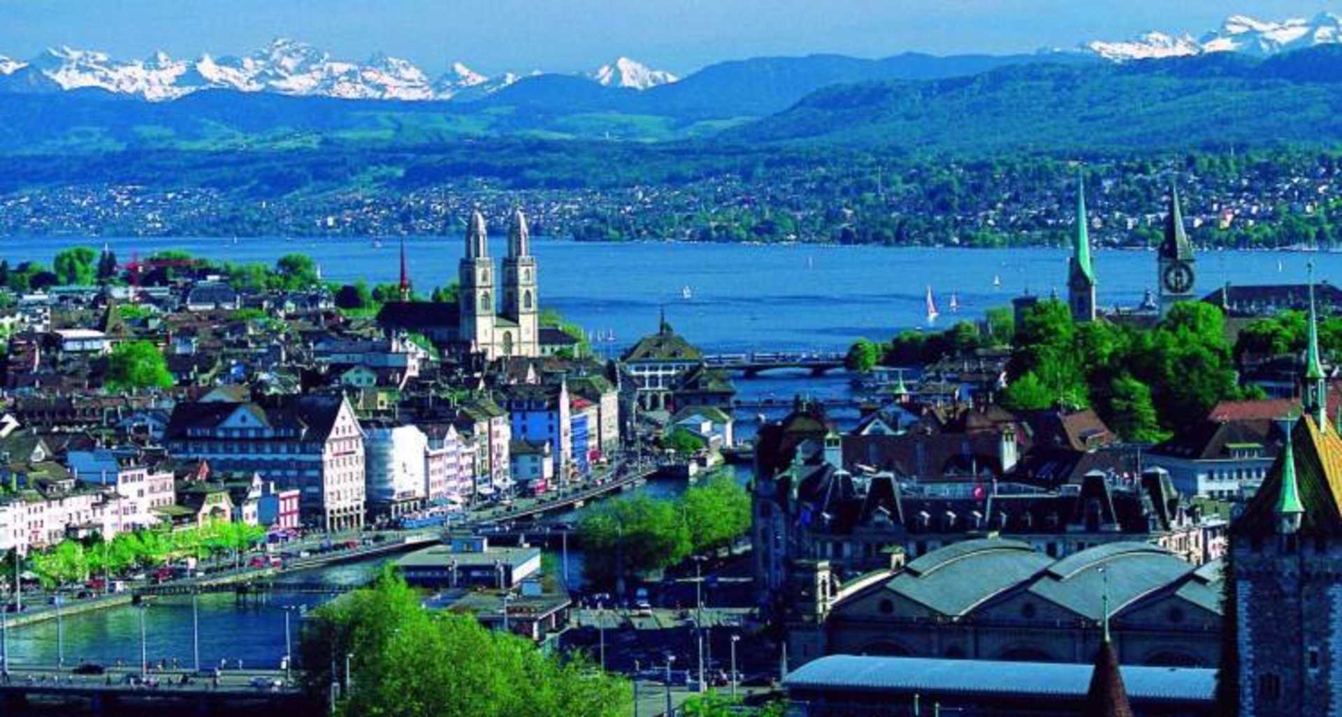 A picturesque city nestled along the shores of a serene lake, surrounded by majestic snow-capped mountains in the distance, with a mix of historic and modern architecture dotting the landscape.