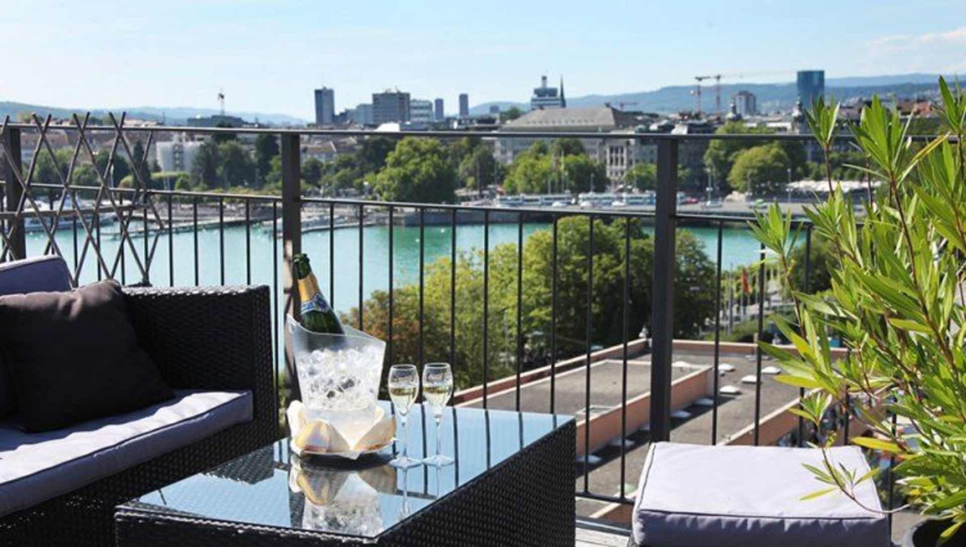 A balcony overlooking a cityscape, with a glass table, wine glasses, and potted plants in the foreground, and a view of high-rise buildings and a river in the background.