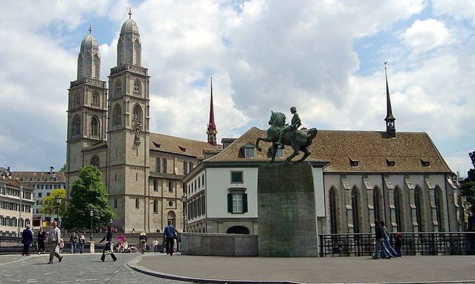 A grand cathedral with tall spires stands in the background, while a statue of a mounted figure is prominently displayed in the foreground. People can be seen walking and cycling around the square.