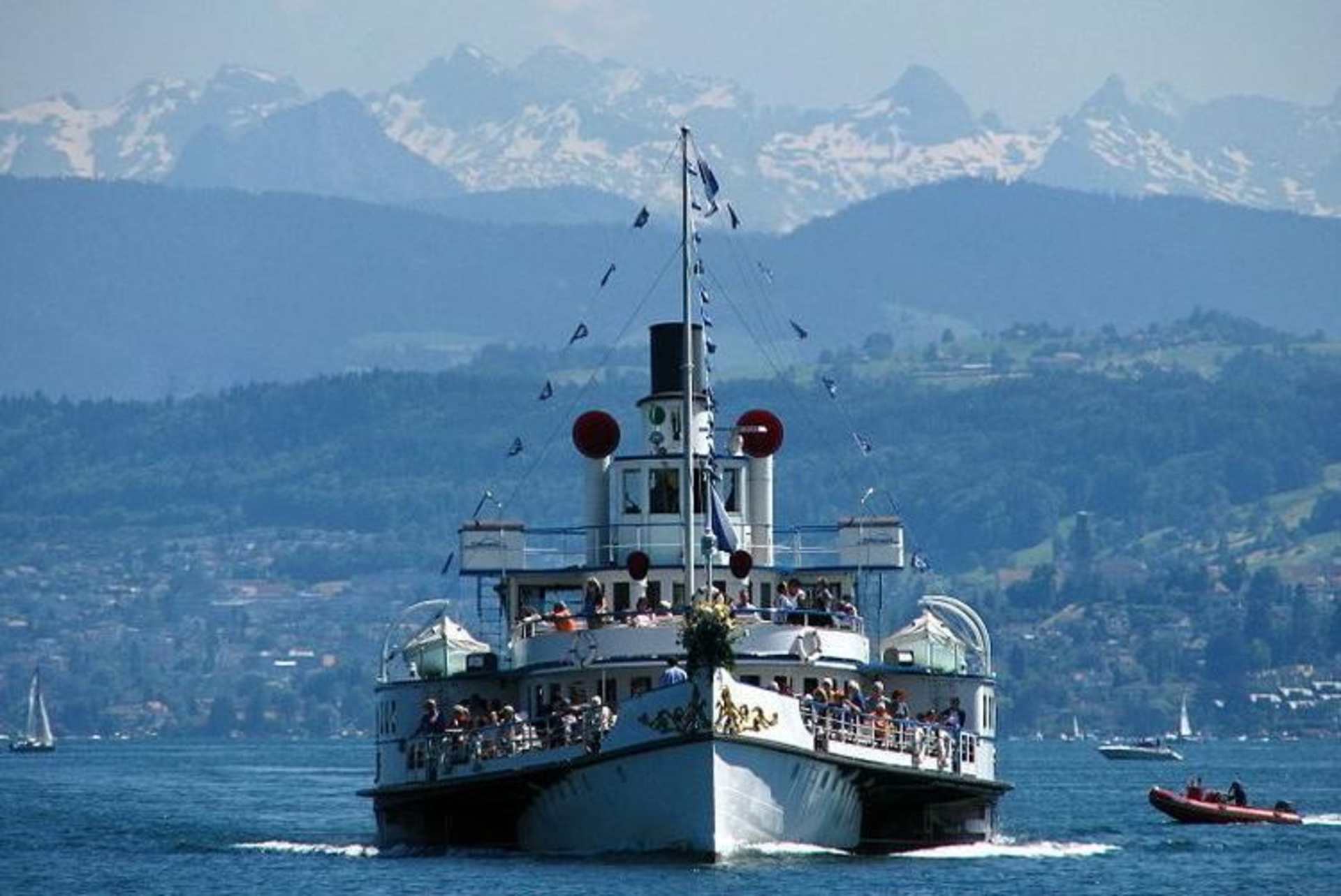 A large passenger ship sails across a scenic lake surrounded by towering snow-capped mountains in the background.