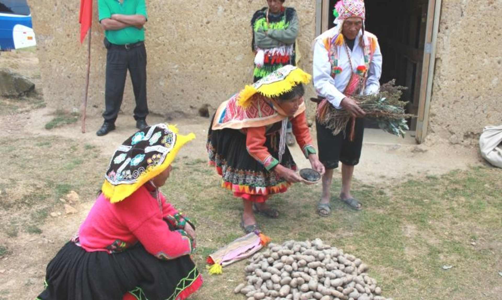The image depicts a group of people in colorful traditional clothing, standing in front of a building and surrounded by various items, including what appears to be a pile of eggs or small round objects.