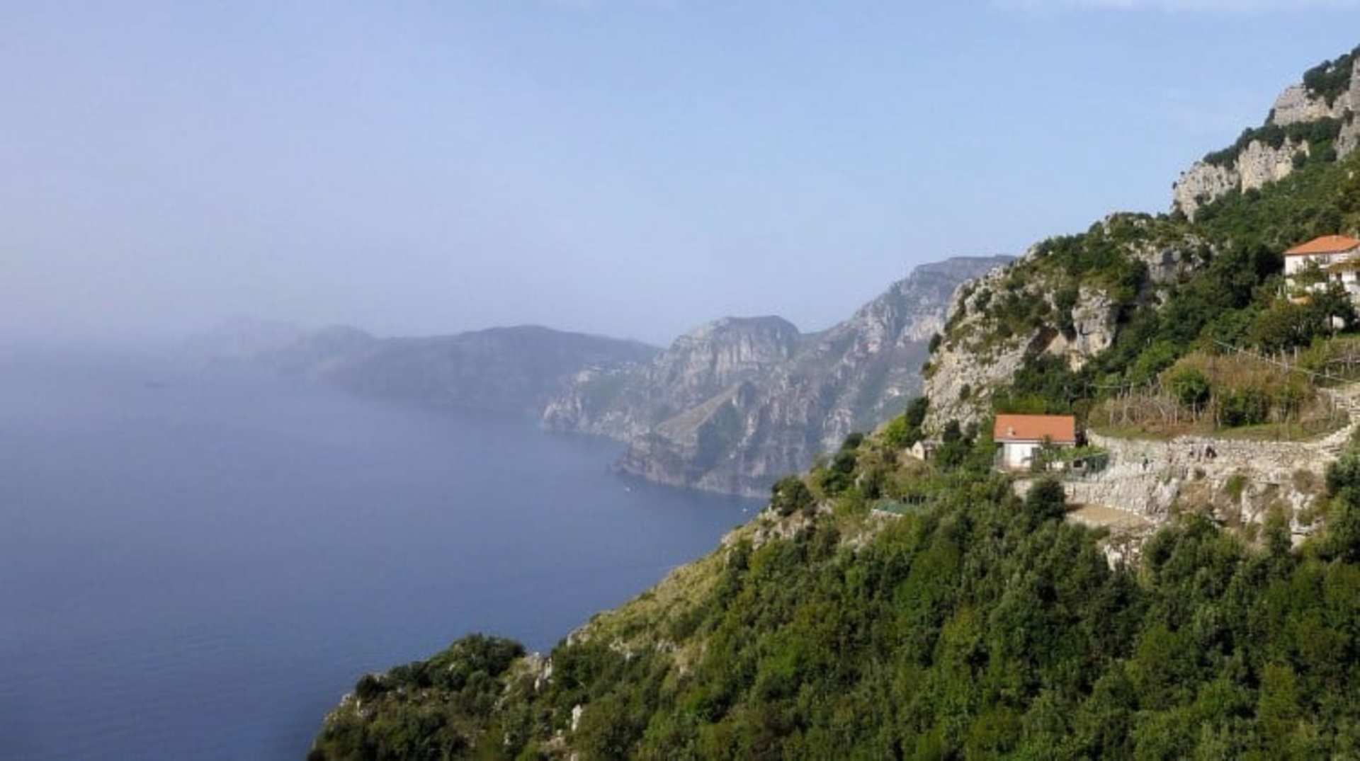 Rugged cliffs and lush greenery line the coastline, with small buildings nestled along the steep slopes overlooking the deep blue waters of the sea.
