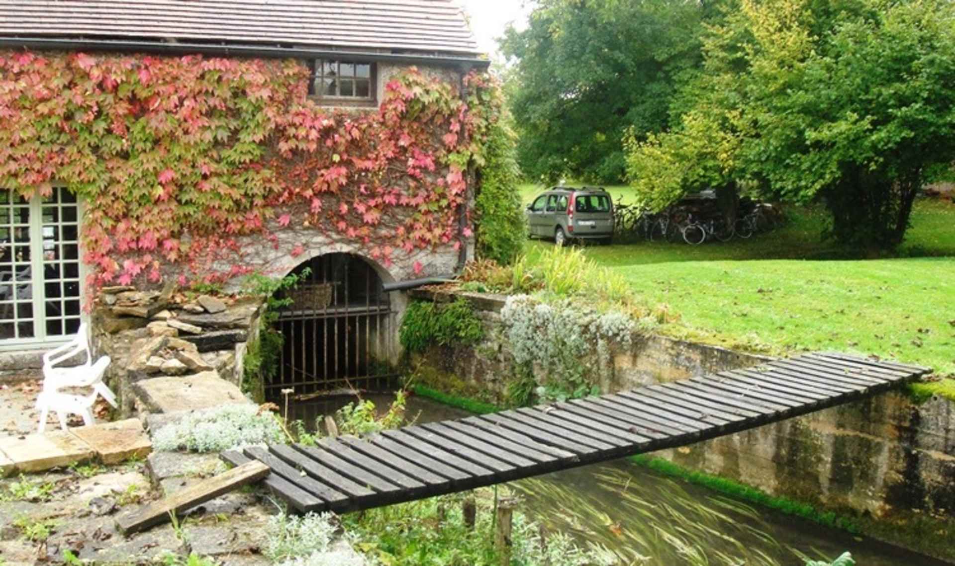 A rustic stone building with a wooden bridge leading to a grassy area, surrounded by lush greenery and a parked vehicle in the background.