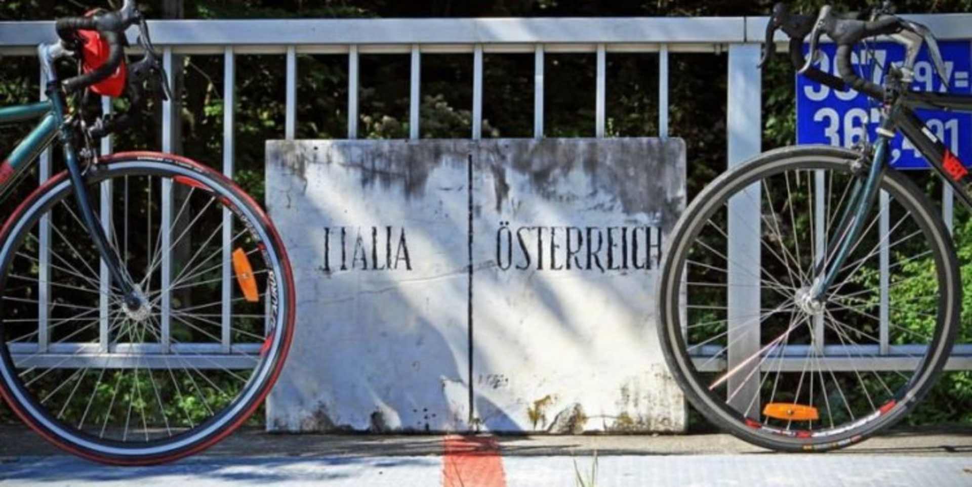 The image shows two bicycles parked in front of a weathered wooden sign that displays the words "Italia Österreich".