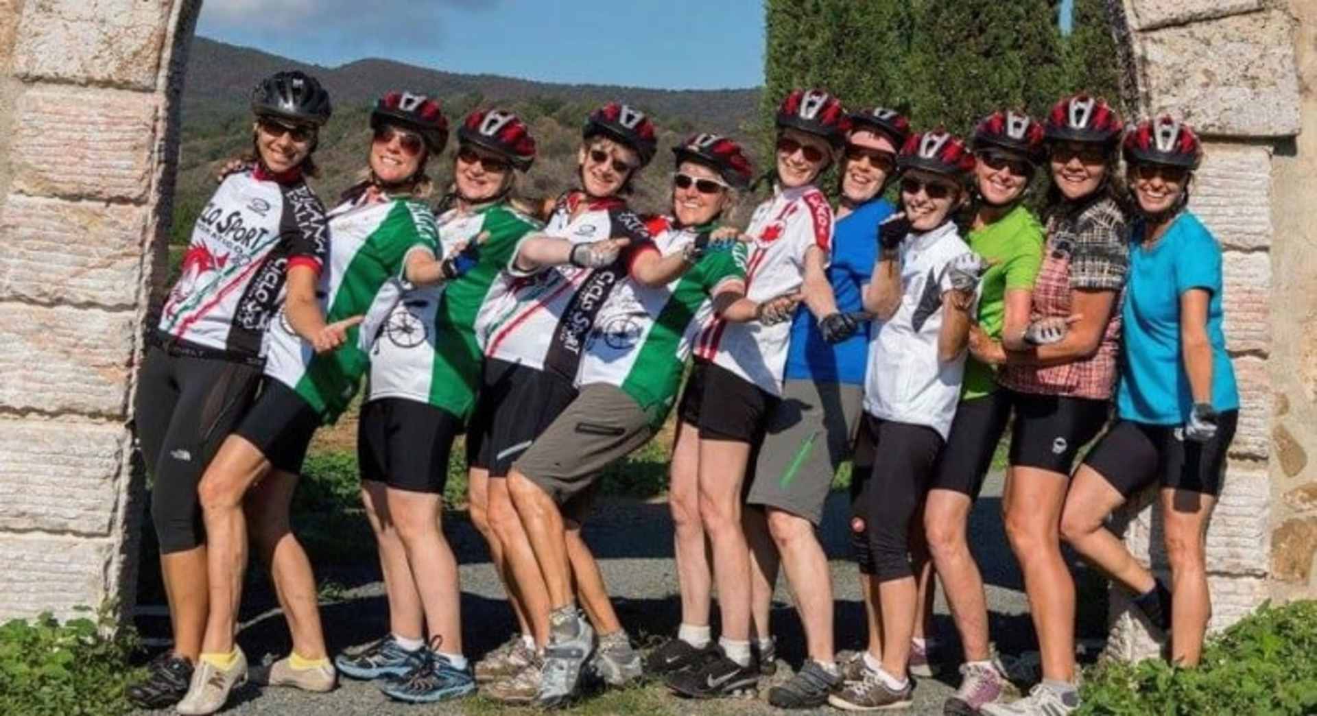 A group of young women in cycling gear posing together in front of a stone wall, with mountains visible in the background.