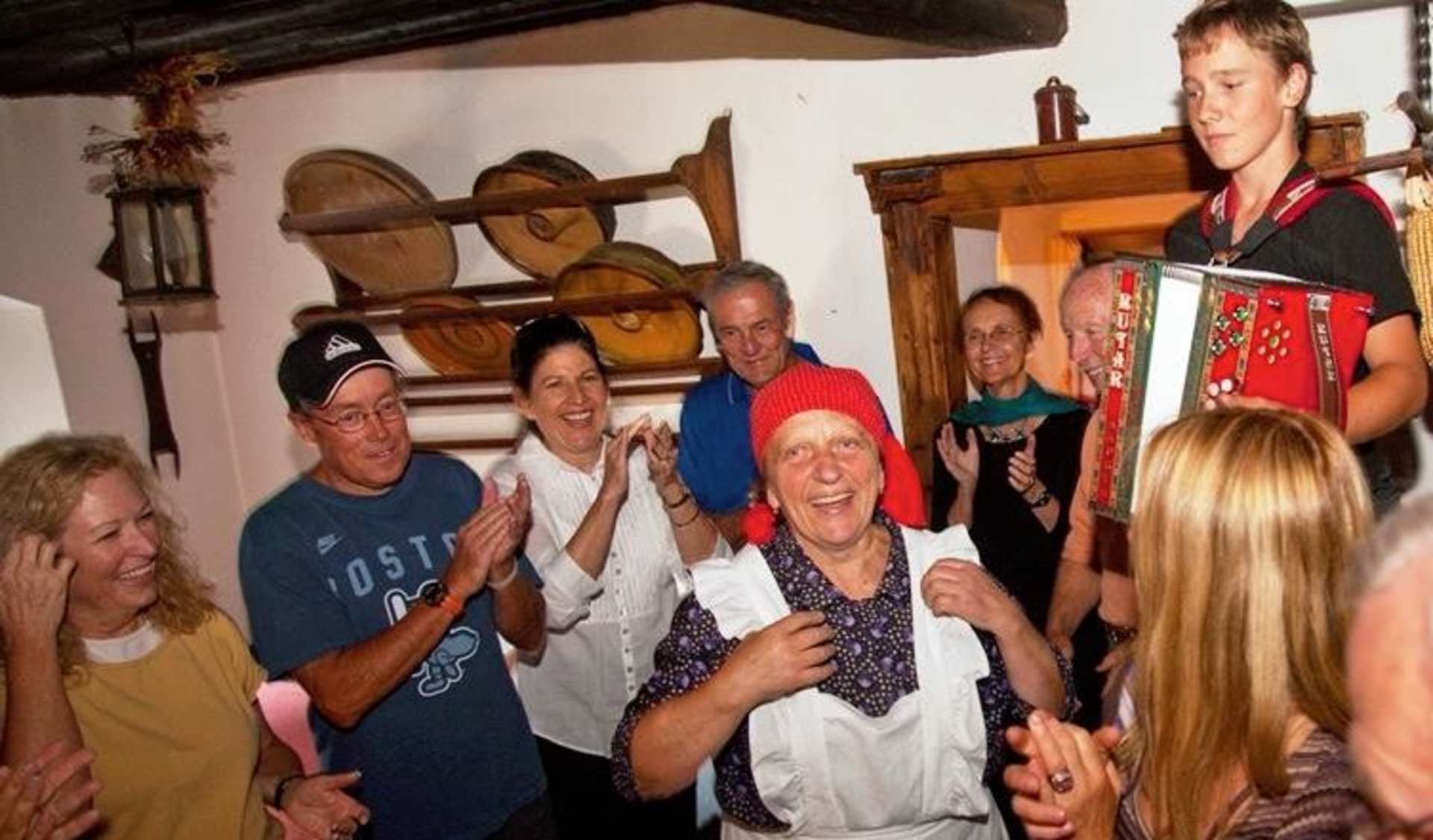 A group of people, both young and old, gathered together in what appears to be a cozy, rustic interior, with wooden shelves and decor in the background.