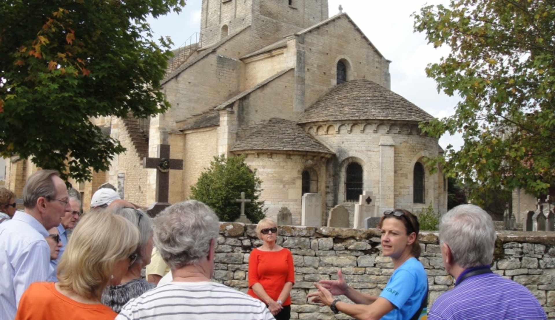 A group of people gathered in front of a historic stone church with a tall steeple, surrounded by trees and a stone wall.