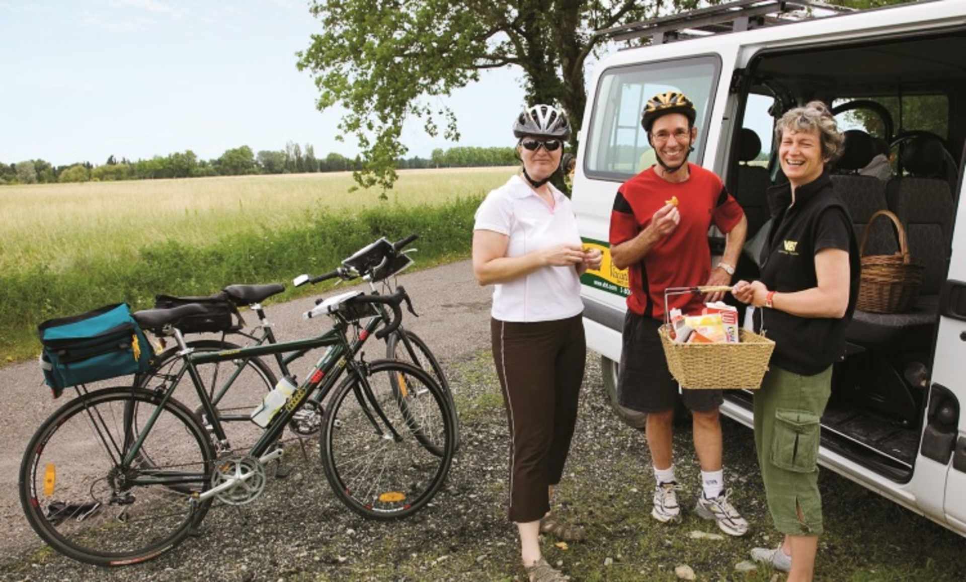 A group of people with bicycles and a van in a rural, grassy field with trees in the background.