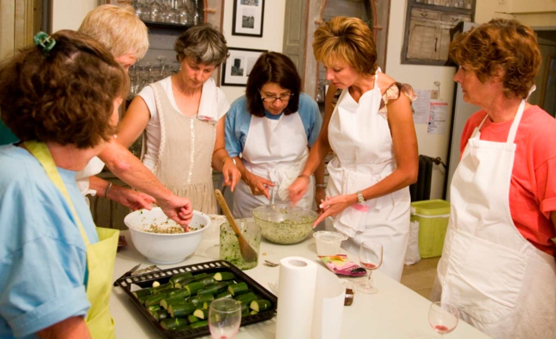 A group of people in aprons are gathered around a table, preparing what appears to be a meal or baking activity together in a cozy, home-like setting with framed artwork on the walls.