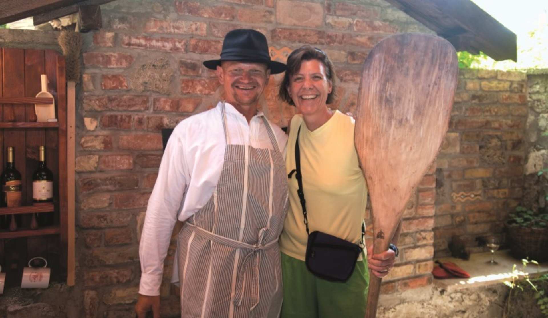 The image shows an older couple standing together in what appears to be a rustic, brick-walled setting, with the man wearing a hat and the woman wearing a yellow top.