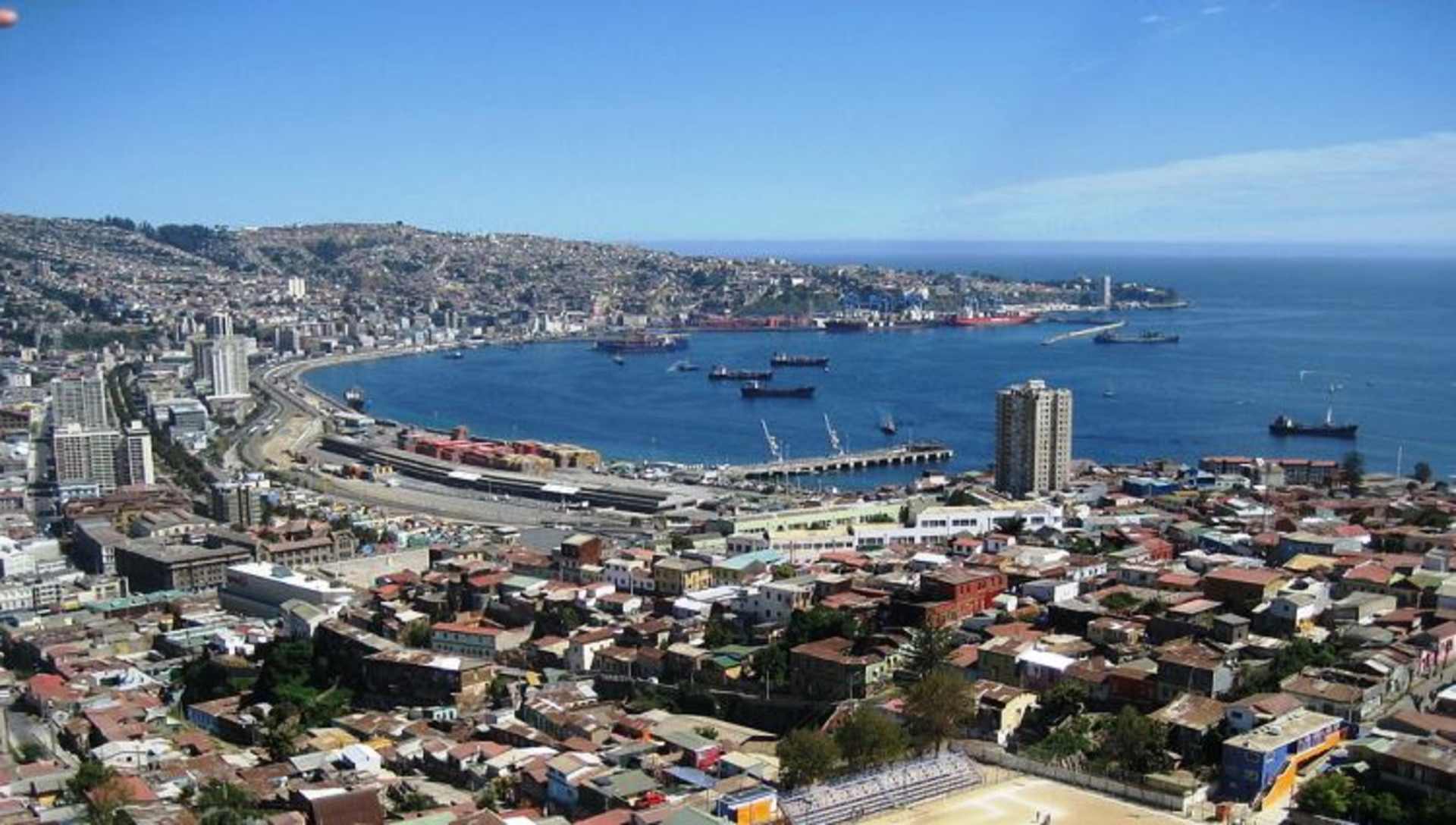 A bustling coastal city nestled between the sea and the hills, with a vibrant harbor filled with ships and boats, surrounded by a dense urban landscape of buildings and infrastructure.