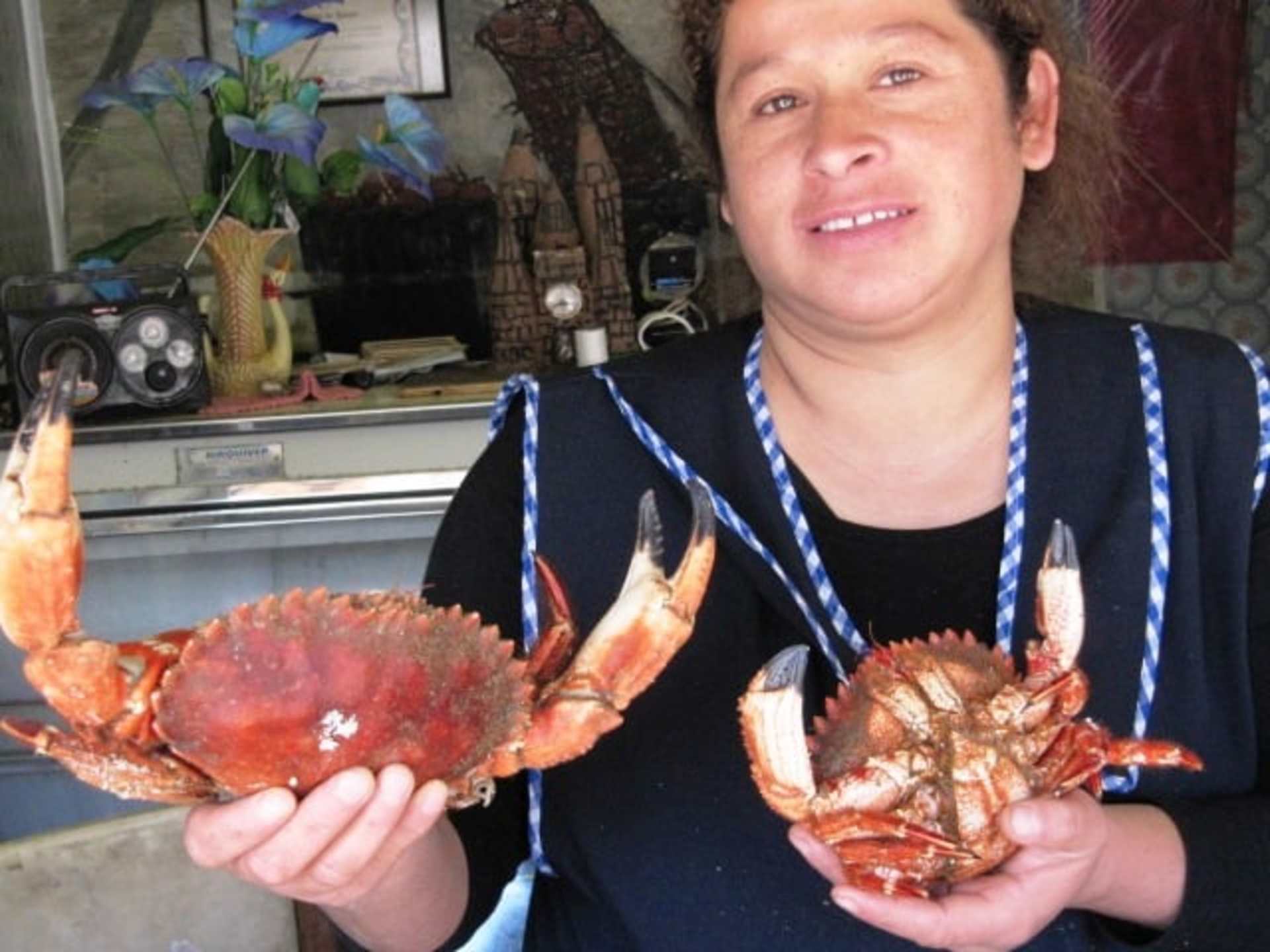 The image shows a person holding two large cooked crabs in their hands, with various kitchen appliances and decorations visible in the background.