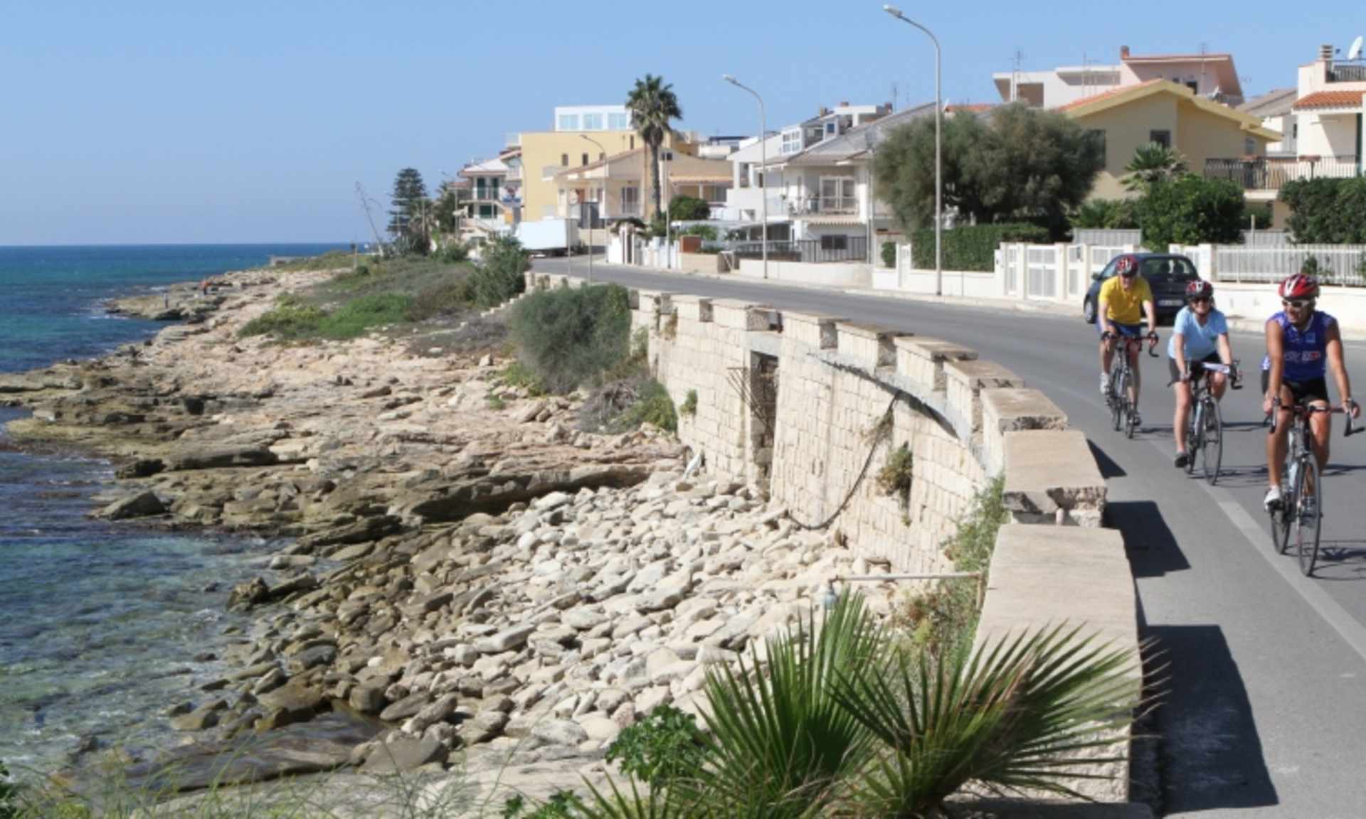 A coastal road lined with colorful buildings and palm trees, with cyclists riding along the rocky shoreline.