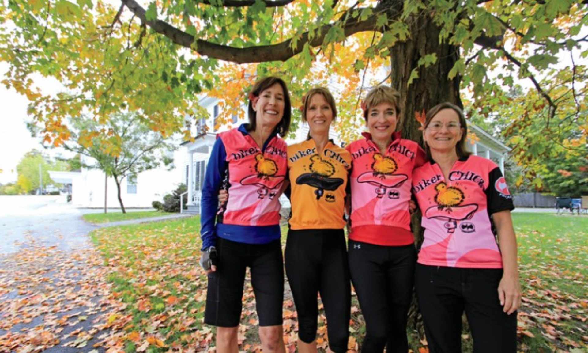 Four women wearing matching pink and orange shirts with a sun design, standing together in an autumn setting with trees and fallen leaves in the background.