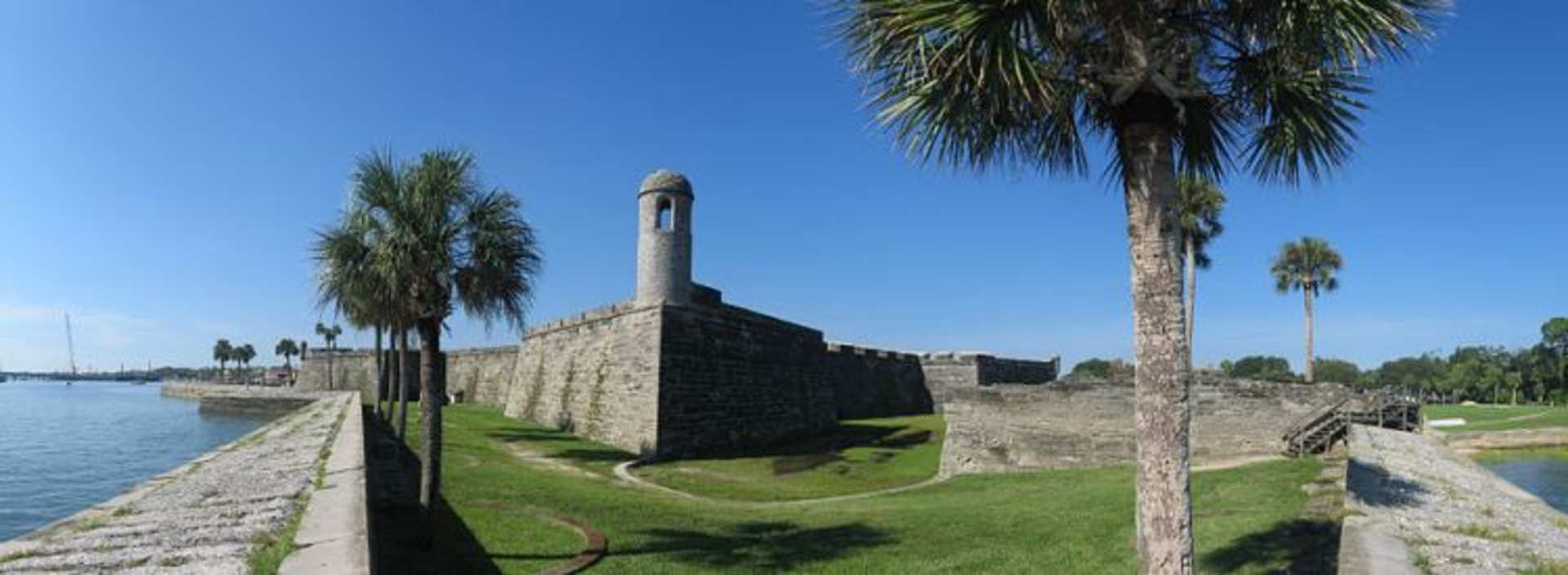 A stone fortress with a tall tower stands amidst lush palm trees, overlooking a grassy area and a body of water in the background.