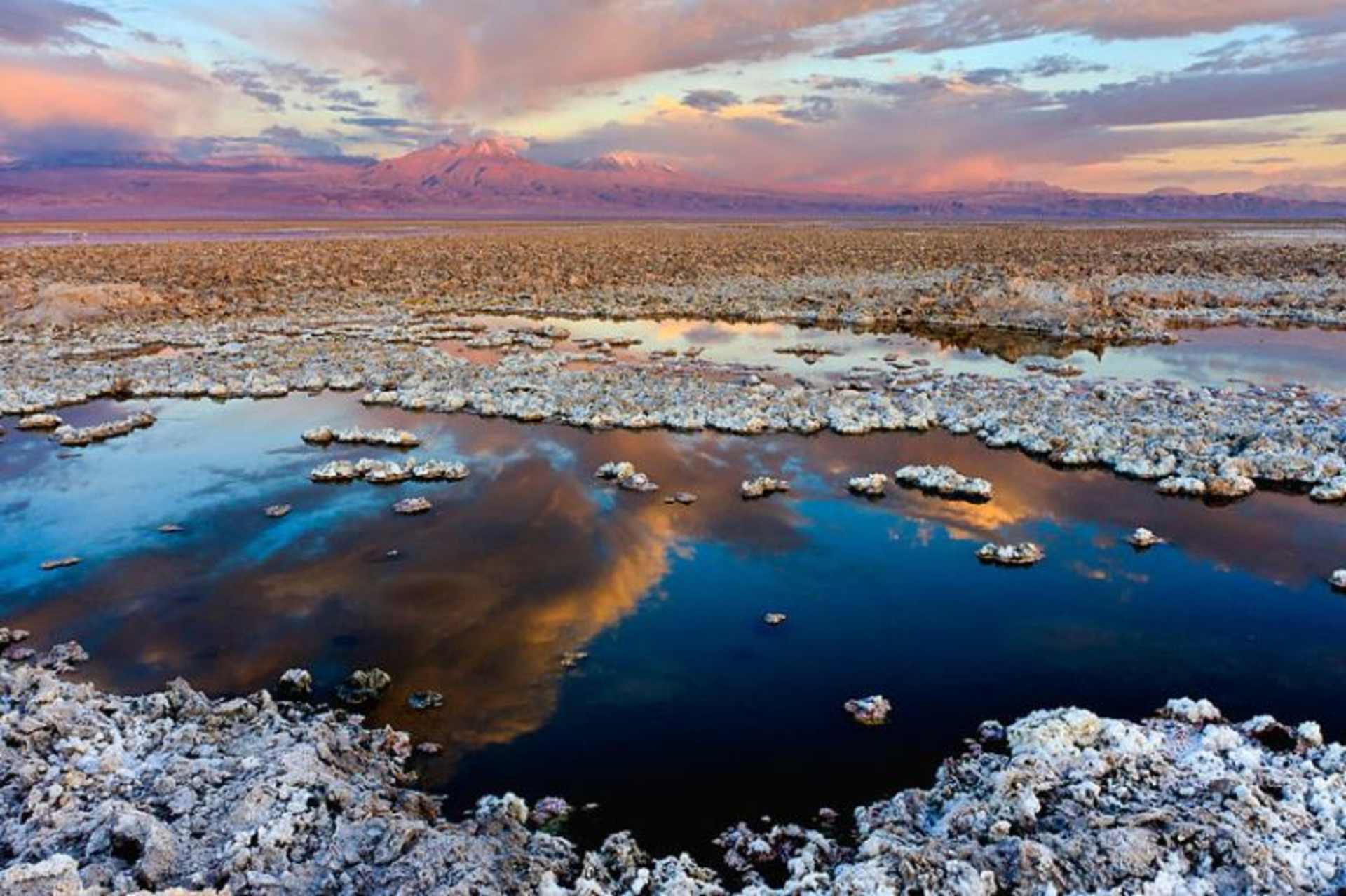 A stunning landscape with a vast, reflective body of water in the foreground, surrounded by snow-covered rocks and a dramatic, colorful sky in the background.