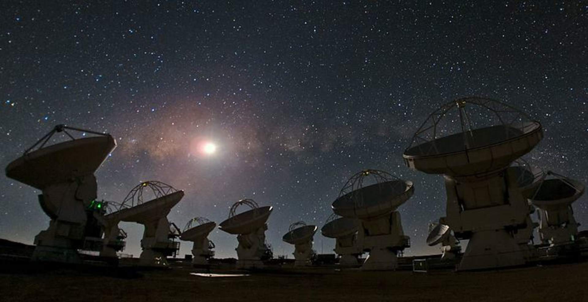 A field of large radio telescopes silhouetted against a starry night sky, with a bright celestial object visible in the background.