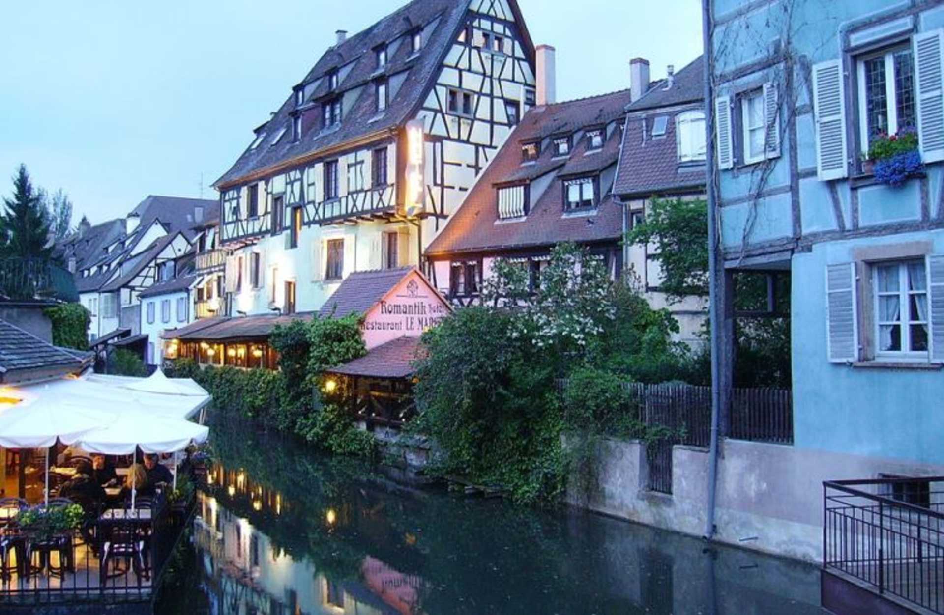 A picturesque canal-side town with charming half-timbered buildings, outdoor cafes, and a tranquil waterway reflecting the colorful architecture.