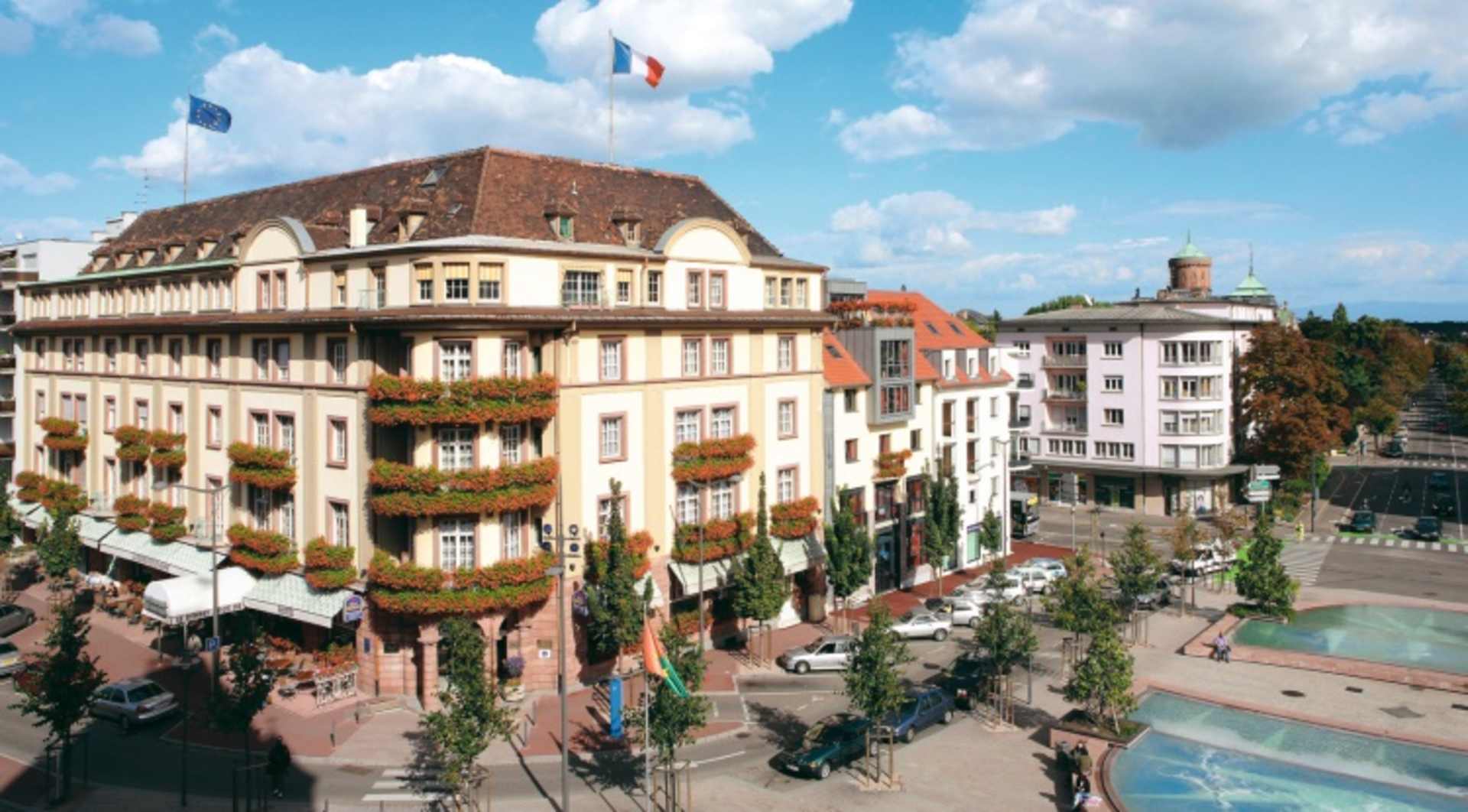 A picturesque European city street lined with ornate, historic buildings, flags, and lush greenery, set against a backdrop of a bright, cloudy sky.