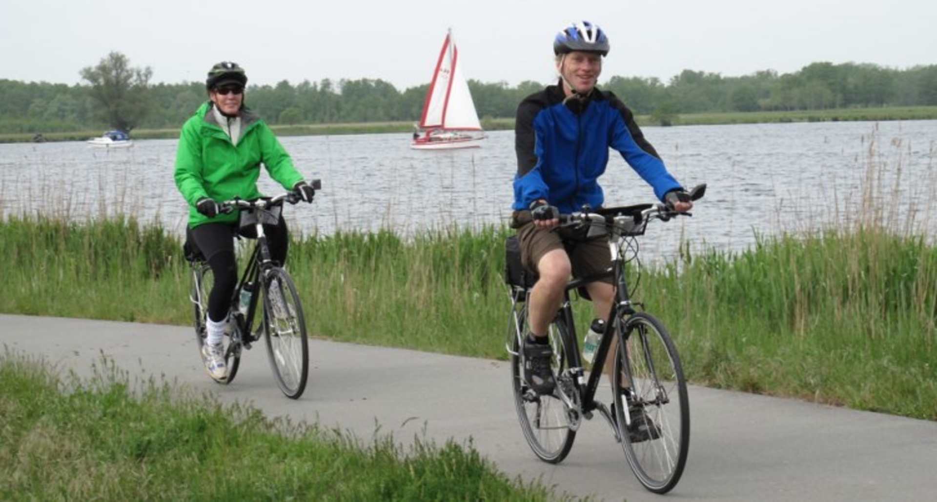Two cyclists, one wearing a green jacket and the other a blue jacket, are riding their bicycles on a path next to a lake with a sailboat in the background.