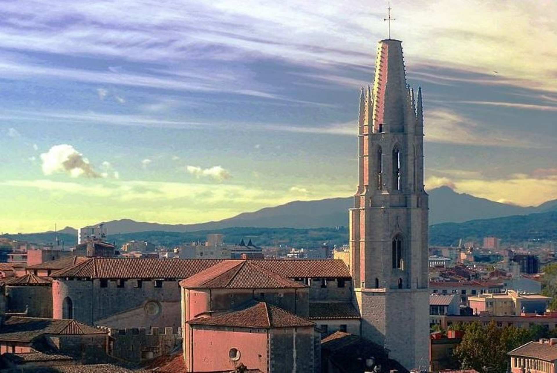 A tall, spire-like tower rises above the rooftops of a city, with mountains visible in the background against a colorful sky.