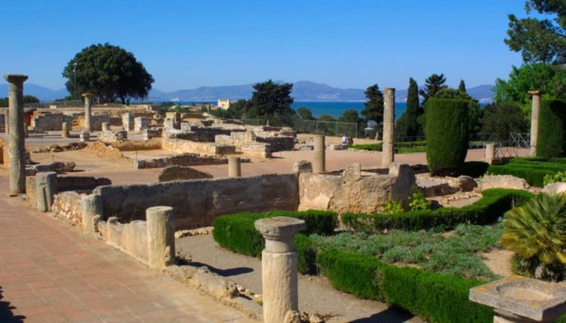 Ruins of an ancient Roman city with columns and stone structures surrounded by lush greenery, with a scenic coastal landscape visible in the background.