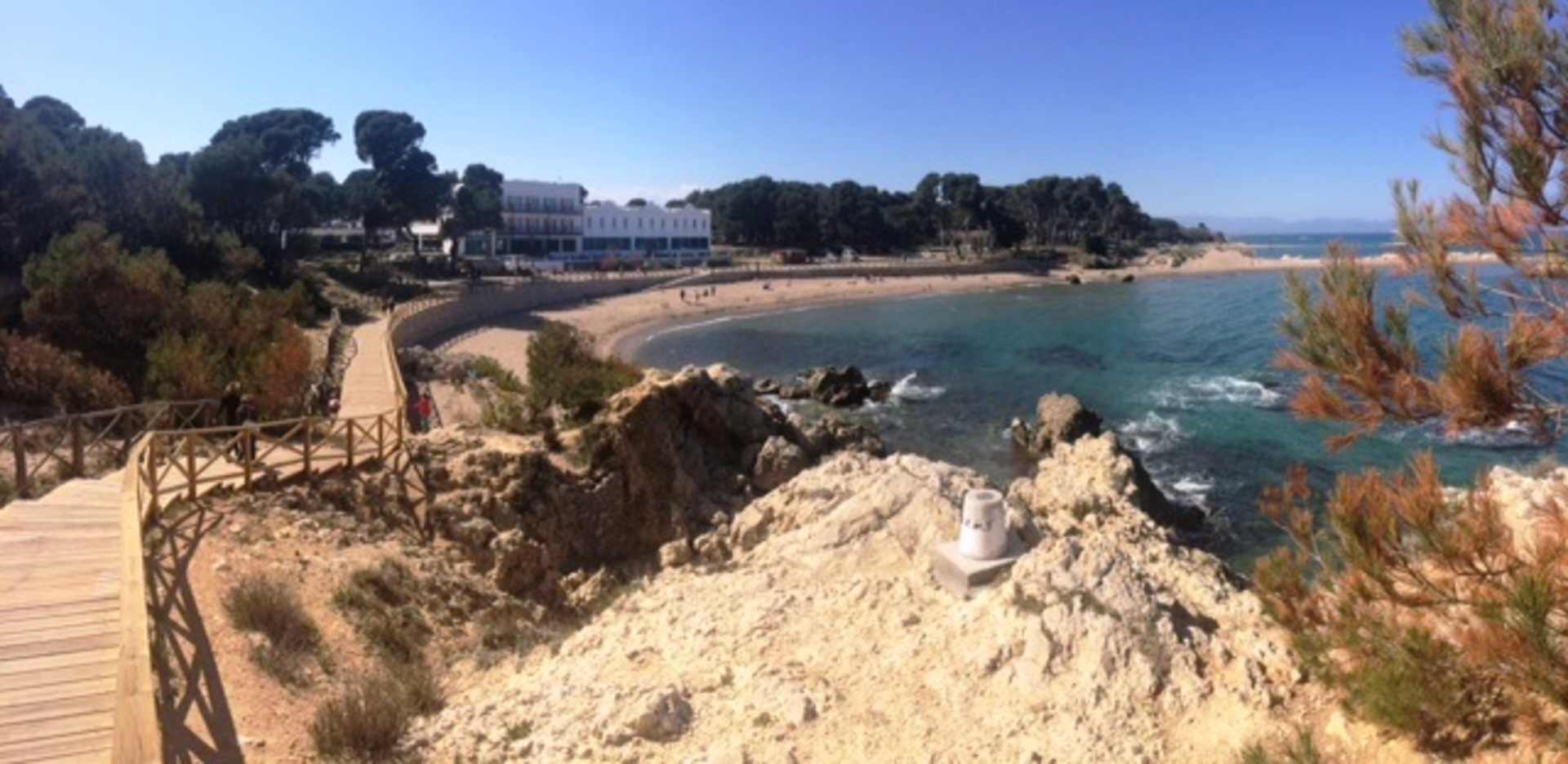 A scenic coastal landscape with a wooden walkway leading along the rocky cliffs, overlooking a turquoise bay and surrounded by lush greenery.