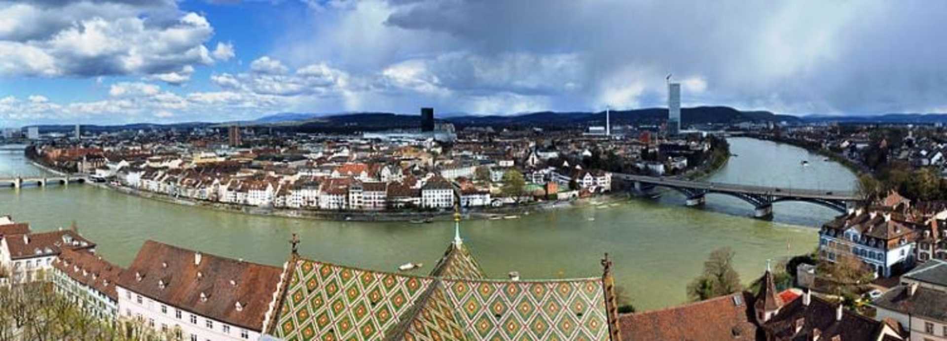 A panoramic view of a picturesque European city nestled along a river, with a mix of historic and modern architecture, surrounded by rolling hills and a vibrant blue sky with fluffy clouds.