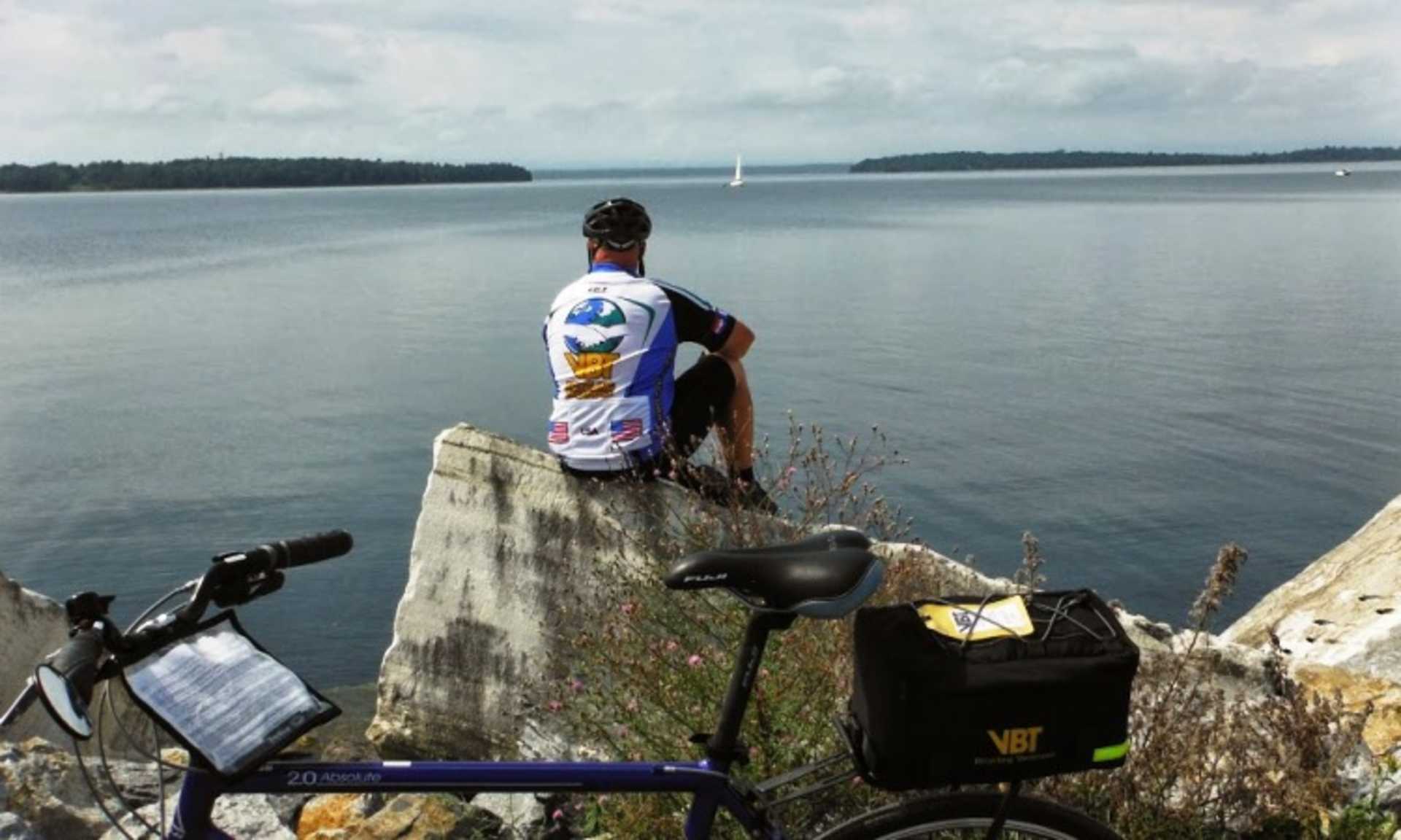 A person stands on a rocky outcrop overlooking a serene lake, with a sailboat visible in the distance.
